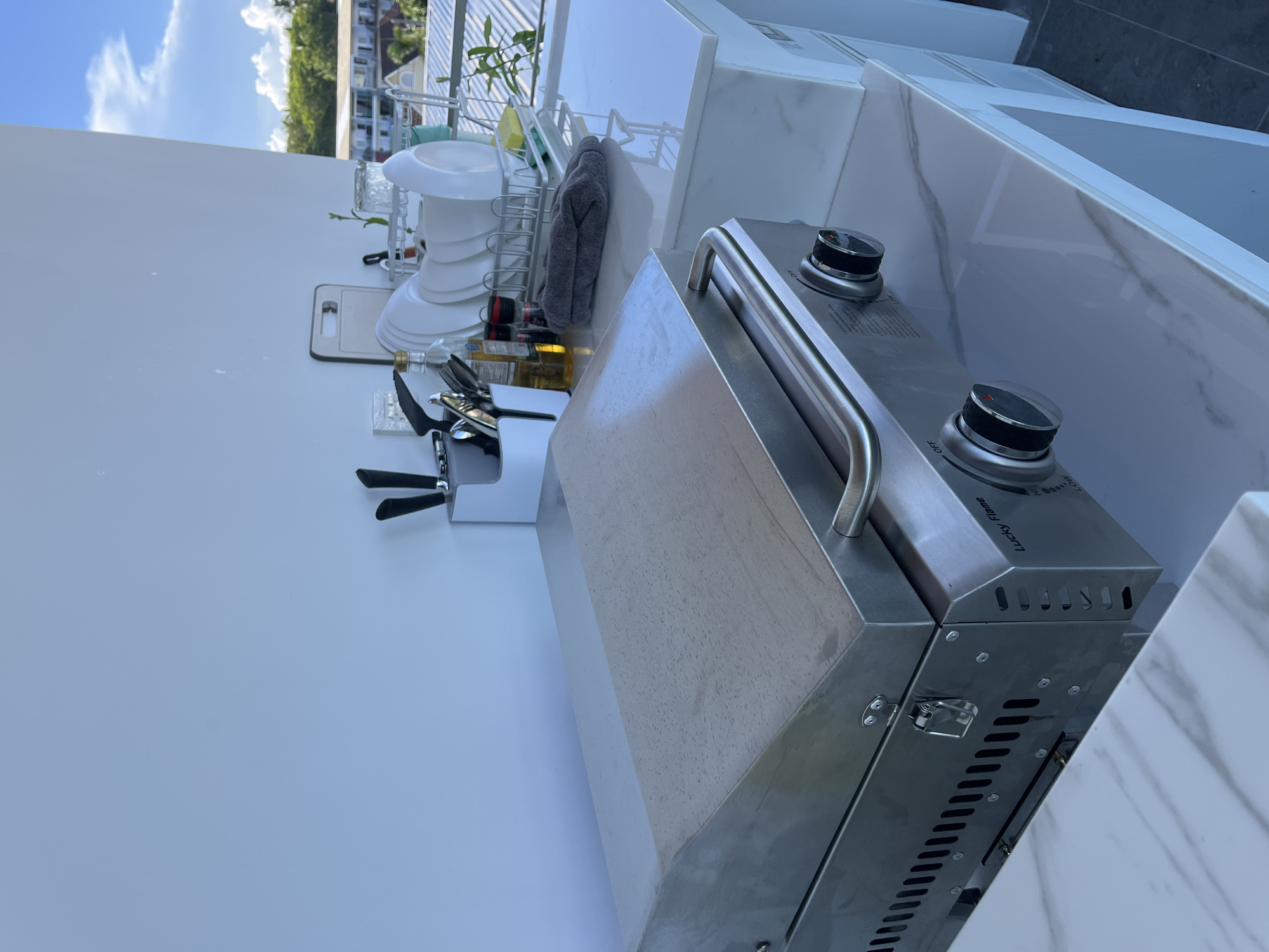 An outdoor cooking area on a balcony in Rawai, Thailand, features a stainless steel griddle with a lid and handles. The griddle is placed on a white marble-patterned countertop, which is part of a modern outdoor kitchen setup. To the right of the griddle, a dish rack holds clean plates, and a cutting board leans against the wall. A utensil holder filled with knives and cooking tools is also visible, along with bottles of oil and seasonings. A dark gray cloth is folded on the countertop near the dish rack. In the background, a lush green landscape with buildings and a bright blue sky with clouds can be seen, suggesting a tropical environment. The overall scene depicts a functional and well-equipped outdoor space.