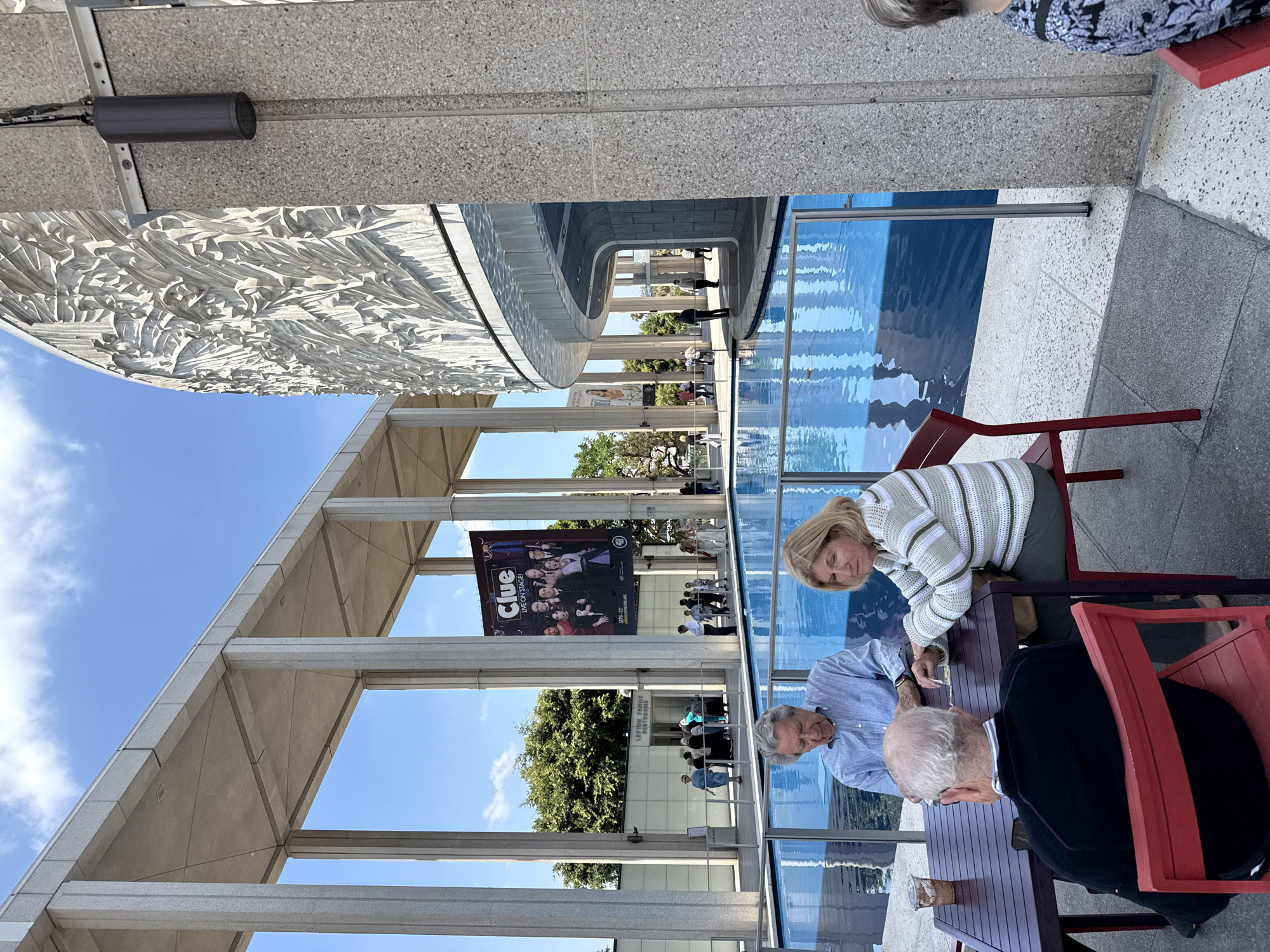 An outdoor daytime shot shows a view of the Music Center in Los Angeles, California. In the foreground, three people are seated at a table next to a blue pool of water. The people appear to be an older couple and an older man. The man on the left is wearing a blue checkered shirt. The woman on the right is wearing a striped sweater. The man in the foreground is wearing a black sweater. The couple is looking down at the table. To the right of the pool is a textured concrete wall with a curvilinear architectural element above it. The wall is lit by a dark cylindrical light fixture. In the background, there is a large banner for the play "Clue" hanging from an overhang supported by concrete pillars. Further in the background, several people are walking near a building labeled "LEFTON FAMILY RESTROOMS". The sky is blue with scattered clouds.