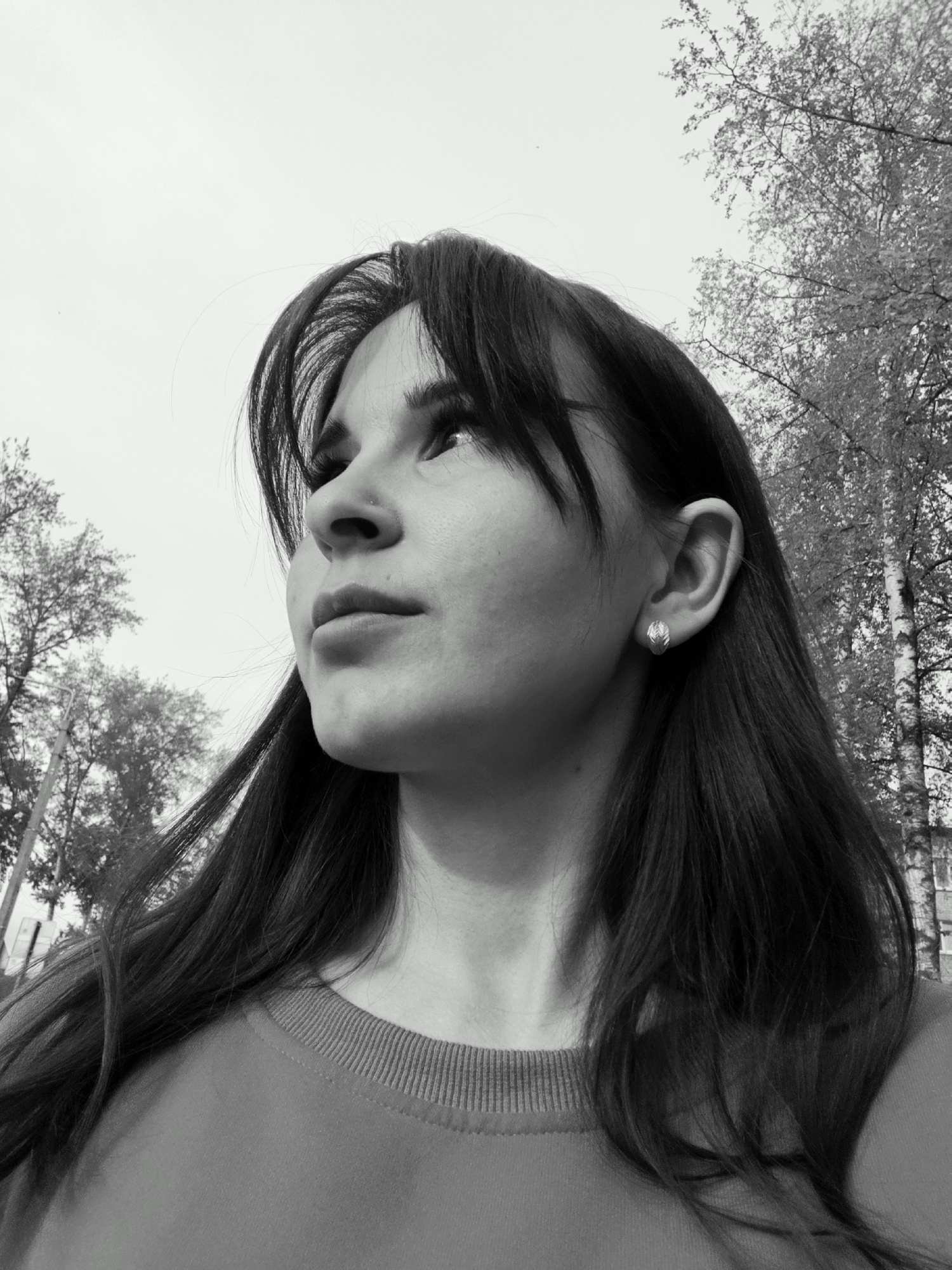 A black and white low-angle close-up shows a woman with long dark hair and bangs looking upward and slightly to the left. She wears earrings and a crew-neck sweater. Behind her are trees, possibly birch, against a light, overcast sky. The location is likely Chaikovsky, Russia, as per the implied context. No specific structures are visible to confirm the exact location within the city.