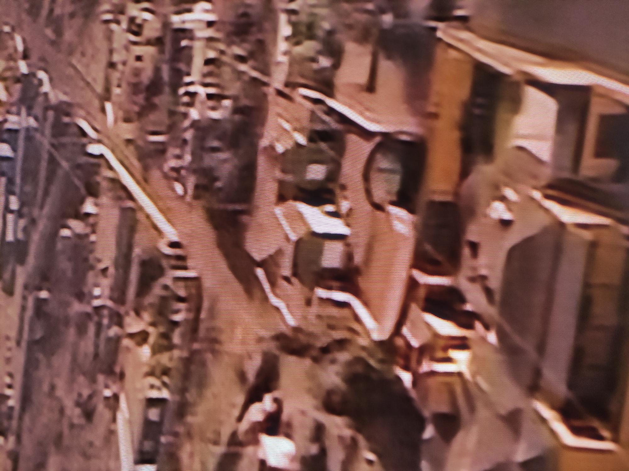 The image and its accompanying crops depict a highly pixelated, blurry, and low-resolution scene, characteristic of a heavily compressed or magnified screen capture, with visible horizontal scan lines.
**Subjects:** The visual information is largely abstract. The scene is dominated by indistinct shapes and forms in muted tones of brown, grey, and dull green. Several prominent lighter, elongated lines traverse the frame, suggesting linear features such as roads, tracks, or boundaries. Interspersed are darker, irregular masses and patches that could be interpreted as clusters of buildings, vegetation, or other structures. Some circular or rounded shapes are faintly visible within these darker areas. While the shapes *could* hint at human activity or vehicles, no identifiable people or specific objects are clearly discernible.
**Activity or event taking place:** Due to the extreme lack of clarity, no specific activity or event can be identified. The composition generally suggests a landscape or an aerial view of an area, possibly urban, industrial, or semi-rural, containing both linear infrastructure and more amorphous features. There is no clear indication of motion, interaction, or any specific narrative.
**Notable details:** The lighting implies daytime, but the precise time of day or prevailing weather conditions cannot be determined from the muted colors and absence of clear shadows or sky details. There are no identifiable landmarks or distinguishing geographical features within the image that could specifically confirm its context as Pavlodar, Kazakhstan. No text is visible in any of the provided media files. The image quality significantly impedes any detailed analysis or confident interpretation.
