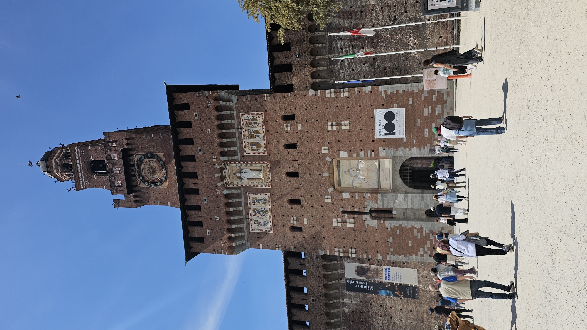 The image shows the Sforza Castle in Milan, Italy. The brick exterior of the castle is the focal point, with the clock tower and the blue sky in the background. There are flags flying from the top of the castle wall. In front of the castle, people are walking along a paved path, casting long shadows. Several banners are visible on the castle wall, including one commemorating Gianfranco Frattini and another advertising an exhibit of Milano and Leonardo.