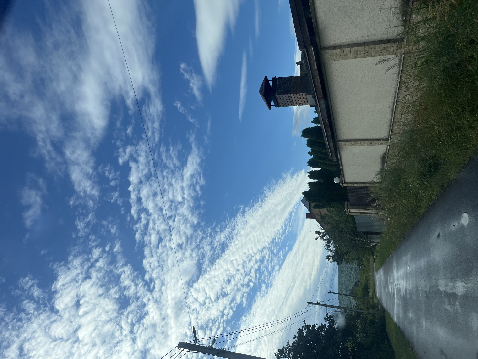 The image shows a view of a paved road in Onokivtsi, Ukraine, bordered by overgrown grass and vegetation on the right and trees and utility poles on the left. The road appears wet, suggesting recent rainfall. In the background, a house with a brick chimney is visible, along with a row of tall, slender trees. The sky is a clear blue with scattered, wispy white clouds, some of which are arranged in parallel bands. The overall impression is a rural or semi-rural setting on a cloudy day. There are no people or animals visible in the image, nor are there any discernible actions or interactions taking place.
