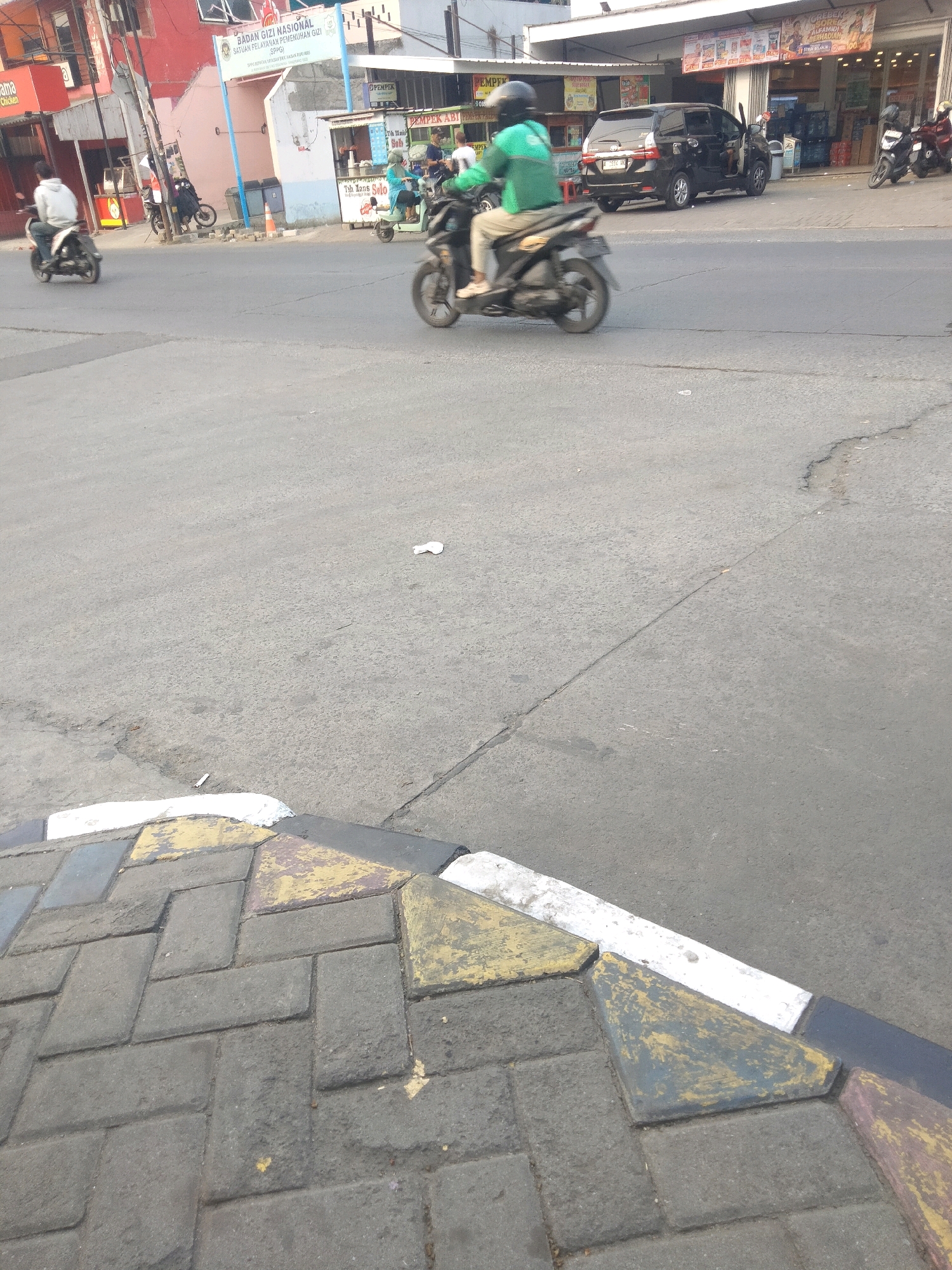 In Sepatan, Indonesia, a street scene captures a section of a road with several motorcycles and a black car. A person on a motorcycle in the foreground wears a green jacket and a helmet, while another person on a motorcycle in the background wears a white hoodie. A black car is parked with its doors open. The road surface is asphalt, with a curb made of interlocking bricks and painted concrete sections. Buildings line the street in the background, featuring signs with text in Indonesian. The lighting suggests it is daytime.