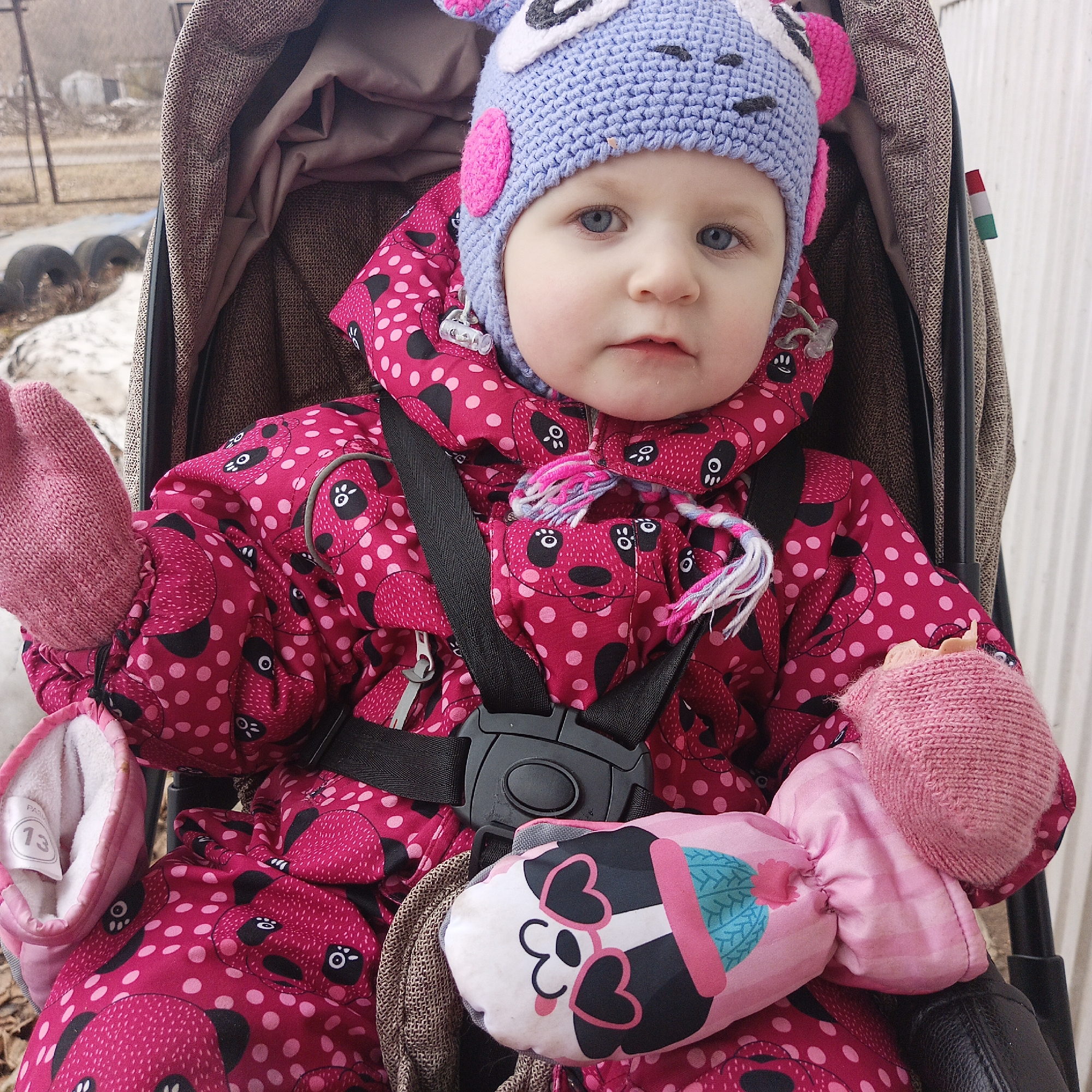 A young child sits in a brown stroller, wearing a pink snowsuit with a black and pink panda bear pattern, a blue crocheted hat with pink ears, and pink mittens. The stroller has a black safety harness fastened around the child. A pink and white stuffed toy rests beside the child. Outside the stroller, glimpses of a snowy or slushy ground, a metal fence, and tires are visible. A small flag with the colors of the Italian flag can be seen on the right side of the stroller. The photograph was taken in the city of Chaikovsky, Russia.