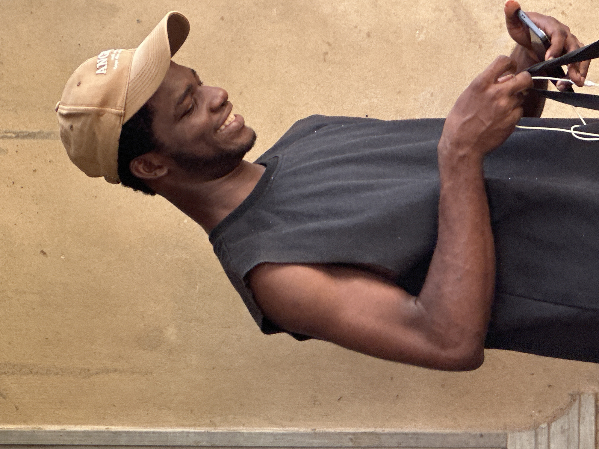 A medium shot depicts a dark-skinned male wearing a tan baseball cap with the word "Angels" embroidered on the front. He is also wearing a sleeveless dark top. The man is smiling and looking down at a cellphone he holds in his hands. White wires are visible near the phone. He appears to be standing outside next to a tan wall. No indication can be found that the image was taken in the city of Ikot Ekpene, Nigeria.