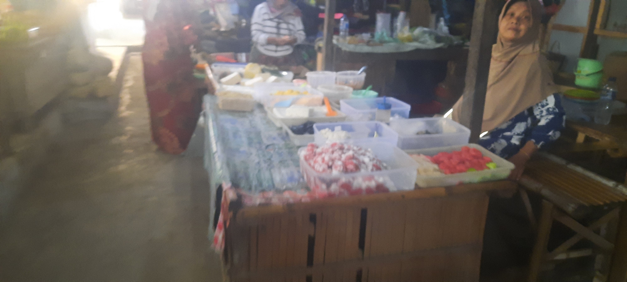 A street-side food stall in Selopuro, Indonesia, is depicted in this underexposed, low-resolution image. The scene features a wooden table laden with various food items in clear plastic containers. These items appear to be colorful sweets or jellies, with visible red and pink assortments. A woman wearing a headscarf and a patterned sarong is seated at the stall, partially obscured by a wooden pillar. In the background, a person in a striped shirt stands behind the table, possibly attending to customers or preparing food. The environment suggests a bustling local market or a roadside vendor setting.