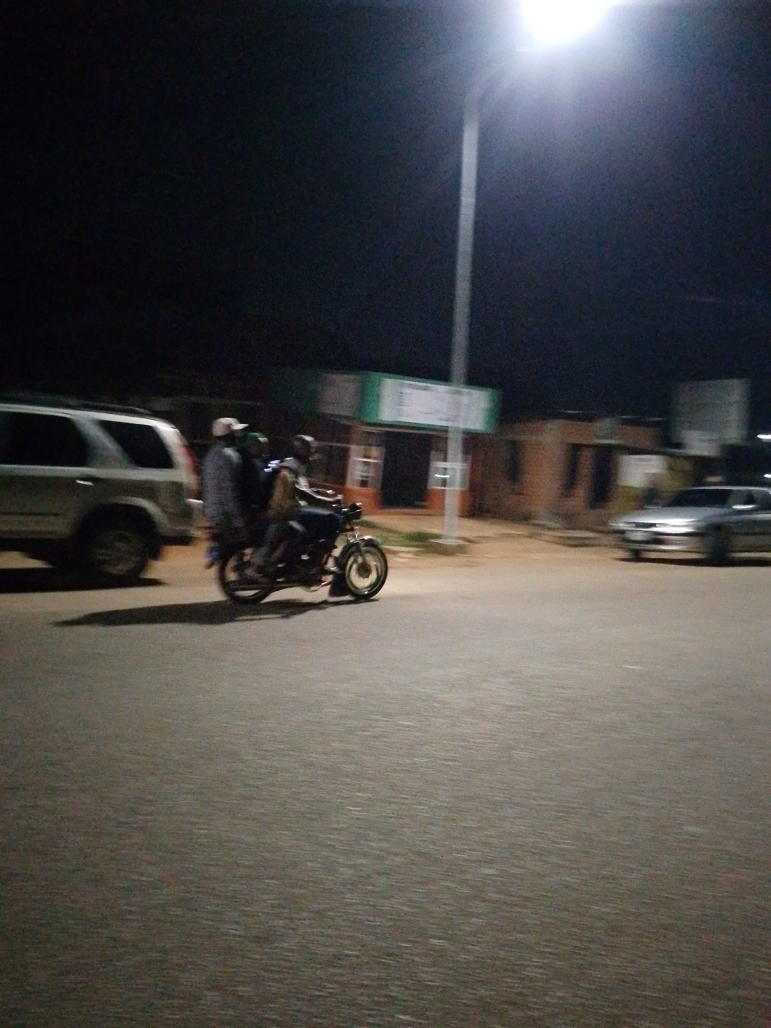 At night, on a paved road in an urban environment, three individuals are riding a motorcycle. The rider is wearing a dark jacket and blue pants. The person in the middle is wearing a white shirt and dark pants, while the person at the back is wearing a light-colored jacket and dark pants. They are positioned in the center of the frame, moving from left to right.
To the left of the motorcycle, a silver SUV is parked. It is a relatively large vehicle with tinted windows. In the background, there are several buildings, including one with a green and white sign. A streetlamp with a bright light is visible in the upper right portion of the image, illuminating the scene. Another silver car is parked in the distance on the right side of the road. The road surface appears to be asphalt, and there is a visible shadow cast by the motorcycle and its passengers. The overall lighting conditions suggest it is nighttime, with artificial light sources being the primary illumination.