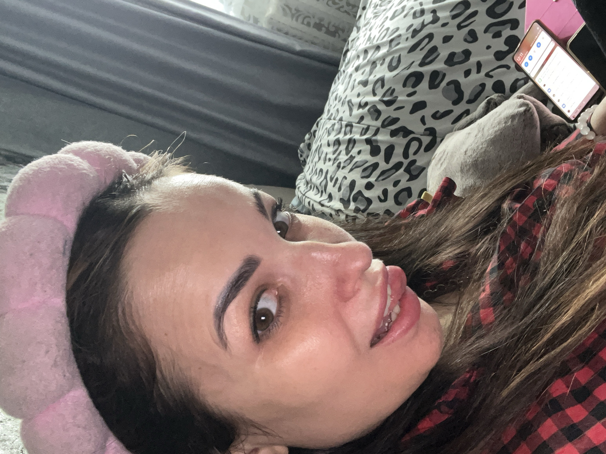 A woman with a pink headband is lying down, looking up and to the left. She has brown hair and brown eyes. She's wearing a red and black plaid shirt. She is holding a phone with a lit screen. Behind her are pillows with animal print and grey fabric. The location is Unknown, Unknown.