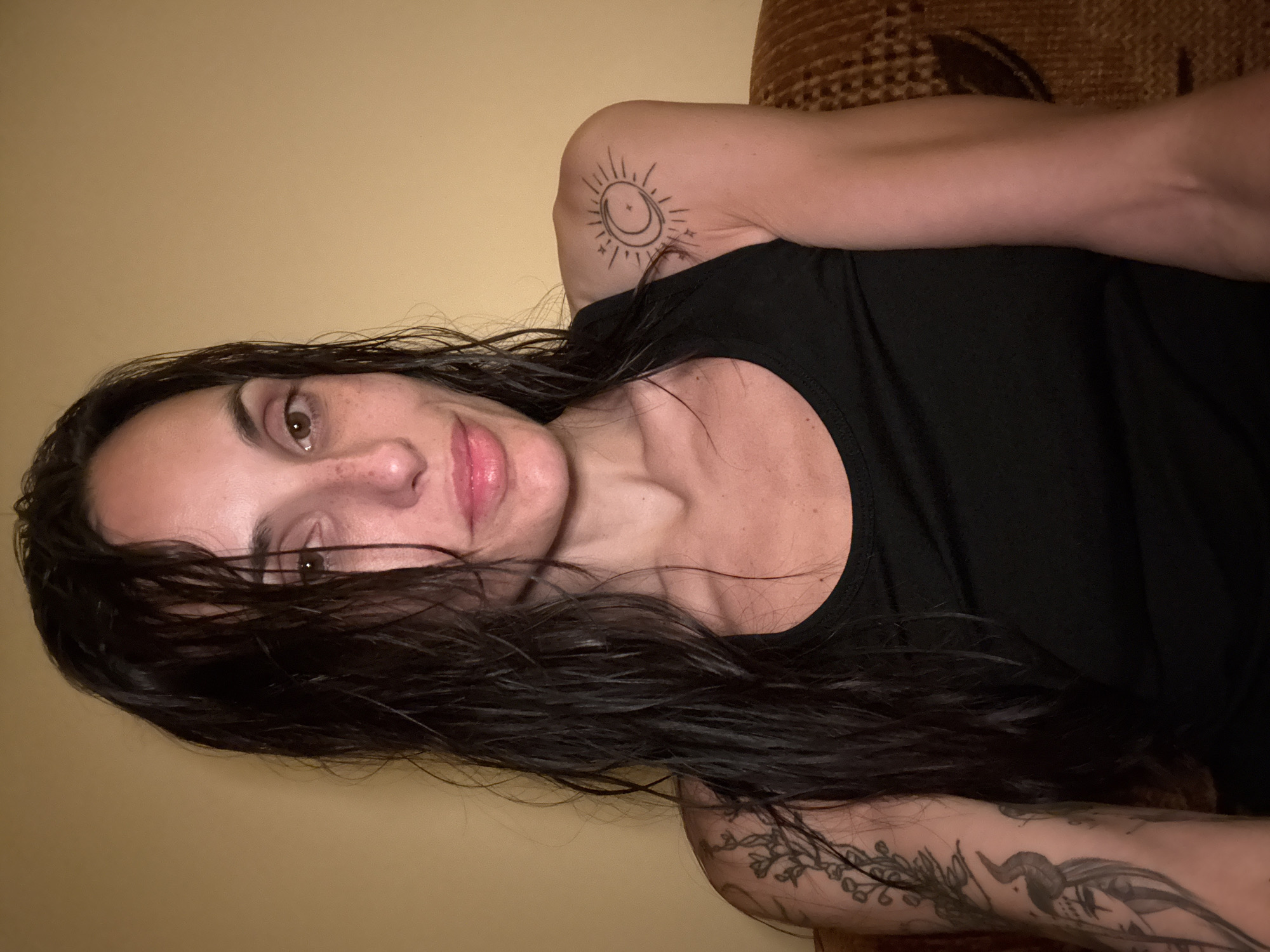 A woman with dark hair and tattoos on her arms is lying down. Her hair is wet and falls across her face and chest. She is wearing a black tank top. The background is a plain beige wall and a brown patterned couch. There is no indication of the location being Pleven, Bulgaria.