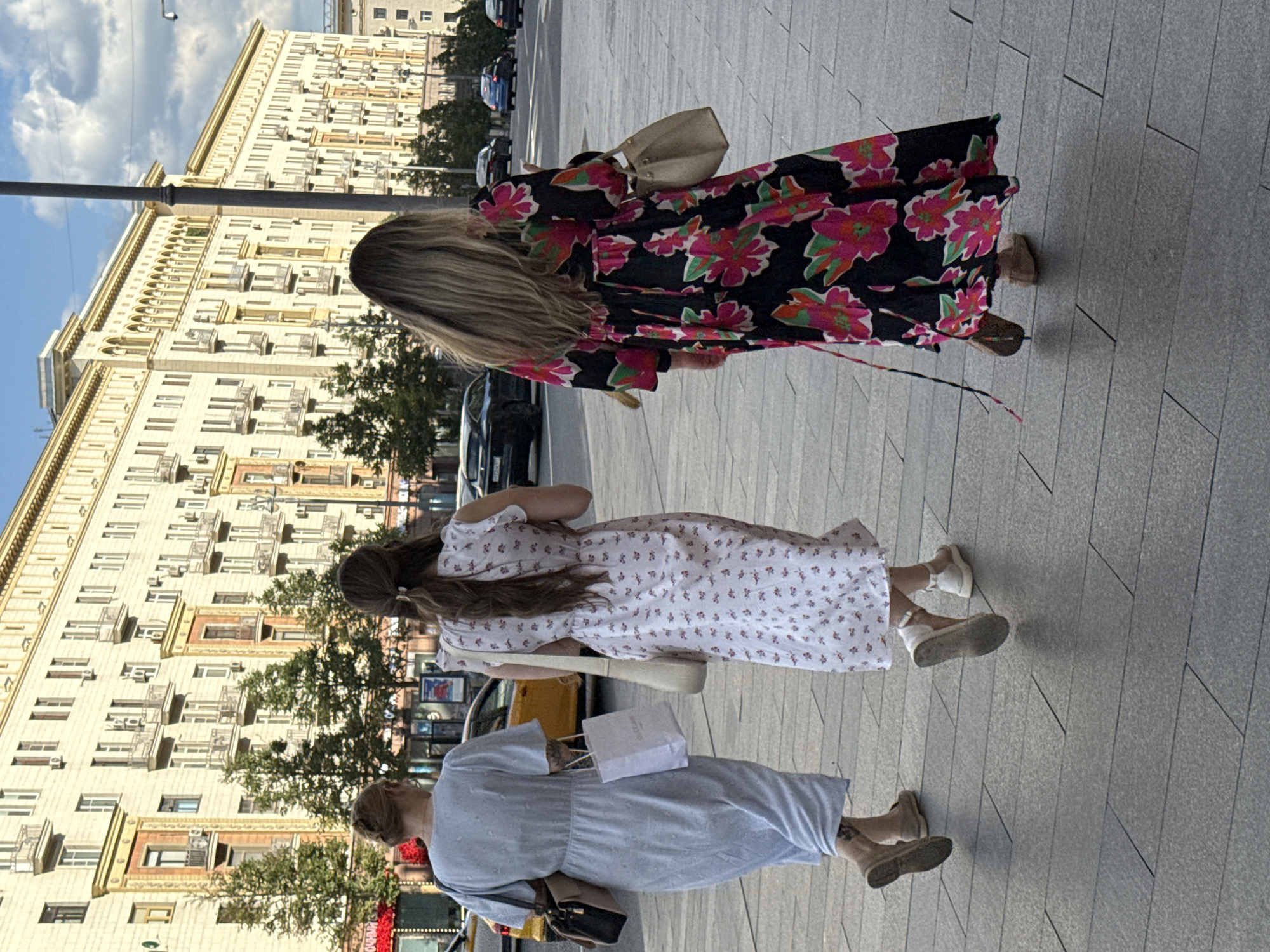 Three individuals are walking away from the camera on a paved sidewalk in front of a large, ornate, multi-story building. The building, with its light-colored facade and numerous windows, appears to be a classic example of Stalinist Empire style architecture. Trees line the sidewalk between the building and a street with cars.
The person on the left is wearing a light blue dress and carrying a black and tan handbag and a white shopping bag. The middle person is wearing a white dress with a small floral pattern and a light-colored crossbody bag. The person on the right is wearing a long, black dress with a bold floral print in shades of pink, fuchsia, and green, and is carrying a light-colored handbag. All three individuals are seen from the waist down, walking in the same direction. The scene suggests a warm day in an urban environment, likely Moscow, Russia, given the architectural style.