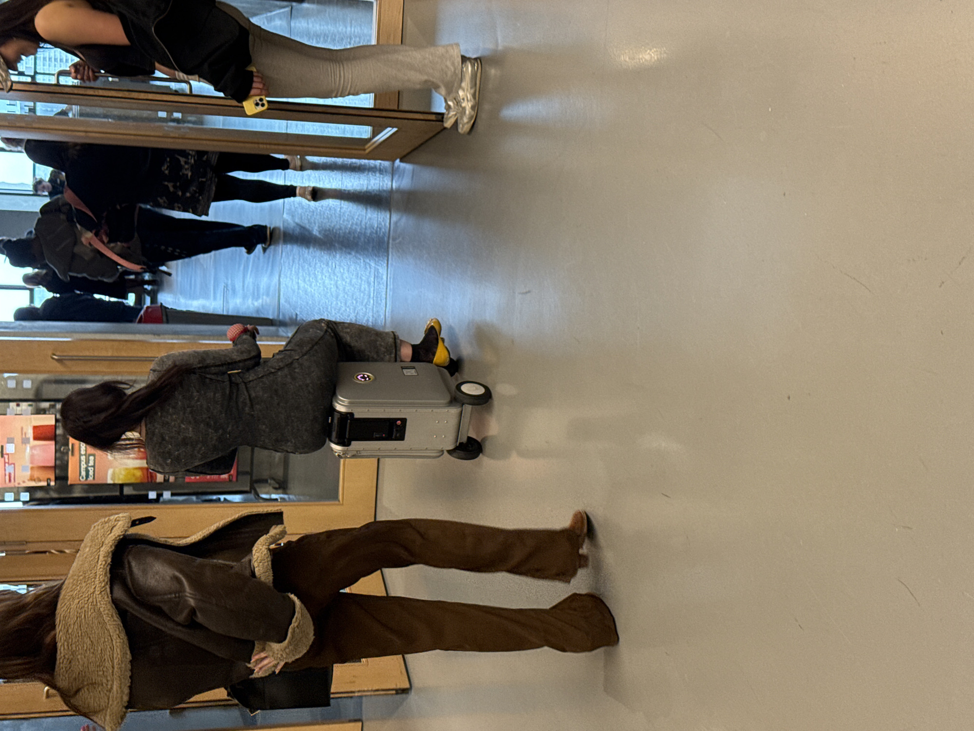 The image shows a number of people entering or exiting a building through a revolving door. One person is seated on a silver, wheeled suitcase, wearing grey clothing and yellow socks. Another person is walking in front of the individual on the suitcase, wearing brown pants and a brown jacket with a light-colored fuzzy lining. Other people are visible entering or exiting through the revolving door. The location is the City of London, United Kingdom, based on the style of the revolving doors and the background details visible through the glass. The setting is indoors, likely a public building or transportation hub, with a smooth, light-colored floor.