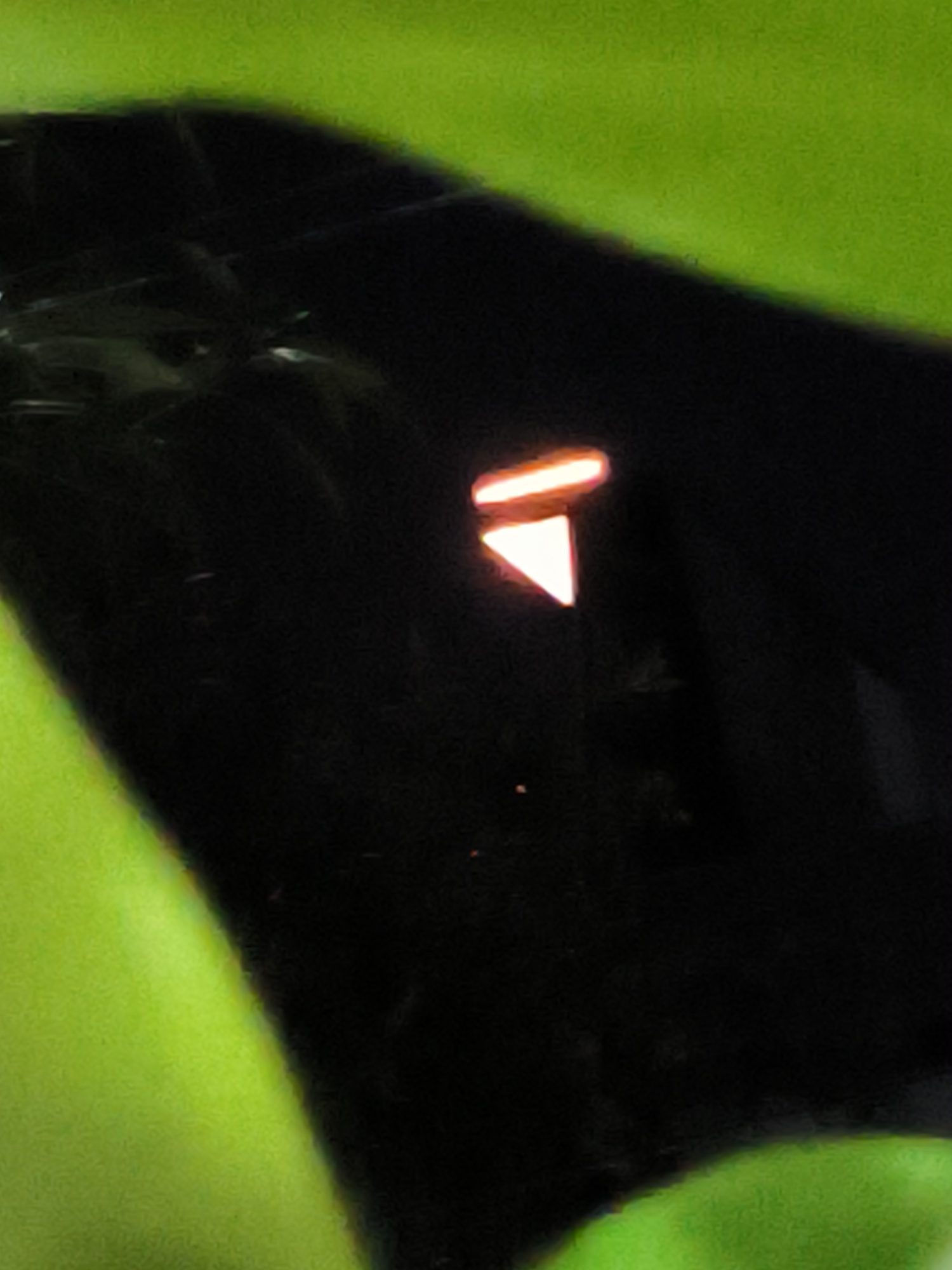 A partially visible, dark and reflective surface is illuminated with a simple graphic. The graphic is a two-part image; a horizontal bar above an inverted triangle. The illuminated graphic is bright orange. The area surrounding the reflective surface is a soft green. There is not enough information to determine its location in the city of Lhokseumawe, Indonesia.