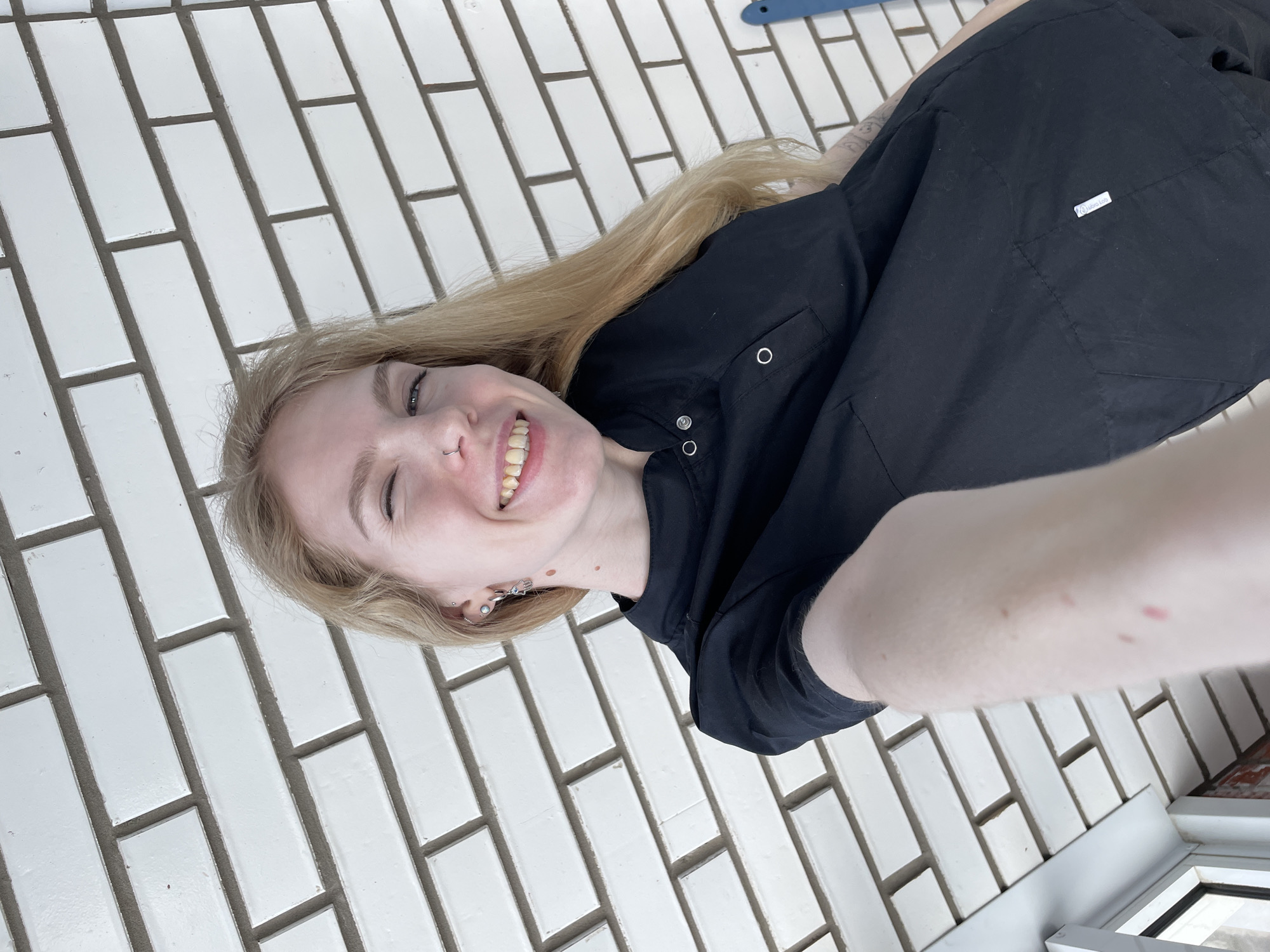 A low-angle, close-up selfie depicts a young adult Caucasian female with long blonde hair, winking her left eye and smiling widely, revealing slightly yellowed teeth. She has a small silver nose ring and multiple silver ear piercings, including a hoop earring with a small charm on her right ear and a stud near the helix. Several small, dark moles are visible on her neck and upper chest. She wears a black collared uniform or scrub top with three visible silver snap buttons on the placket and a small rectangular pocket on the left chest. A white tag with "Just_Job" in black text is affixed to the lower right side of her garment. Faint tattoo markings are visible on her left arm. Red, indistinct marks are present on her right forearm, which extends into the frame, suggesting it is holding the camera. The background consists primarily of a white brick wall with grey mortar, laid in a stretcher bond pattern. In the lower left corner, a white window frame or architectural trim is visible, with a small section of reddish-brown brickwork below it. A blue object, indistinct in form, is partially visible on the far right edge of the frame. No specific landmarks or unique identifiers within the image can confirm a precise location within Izhevsk, Russia; the environment is generic and consistent with urban or suburban residential or commercial settings.