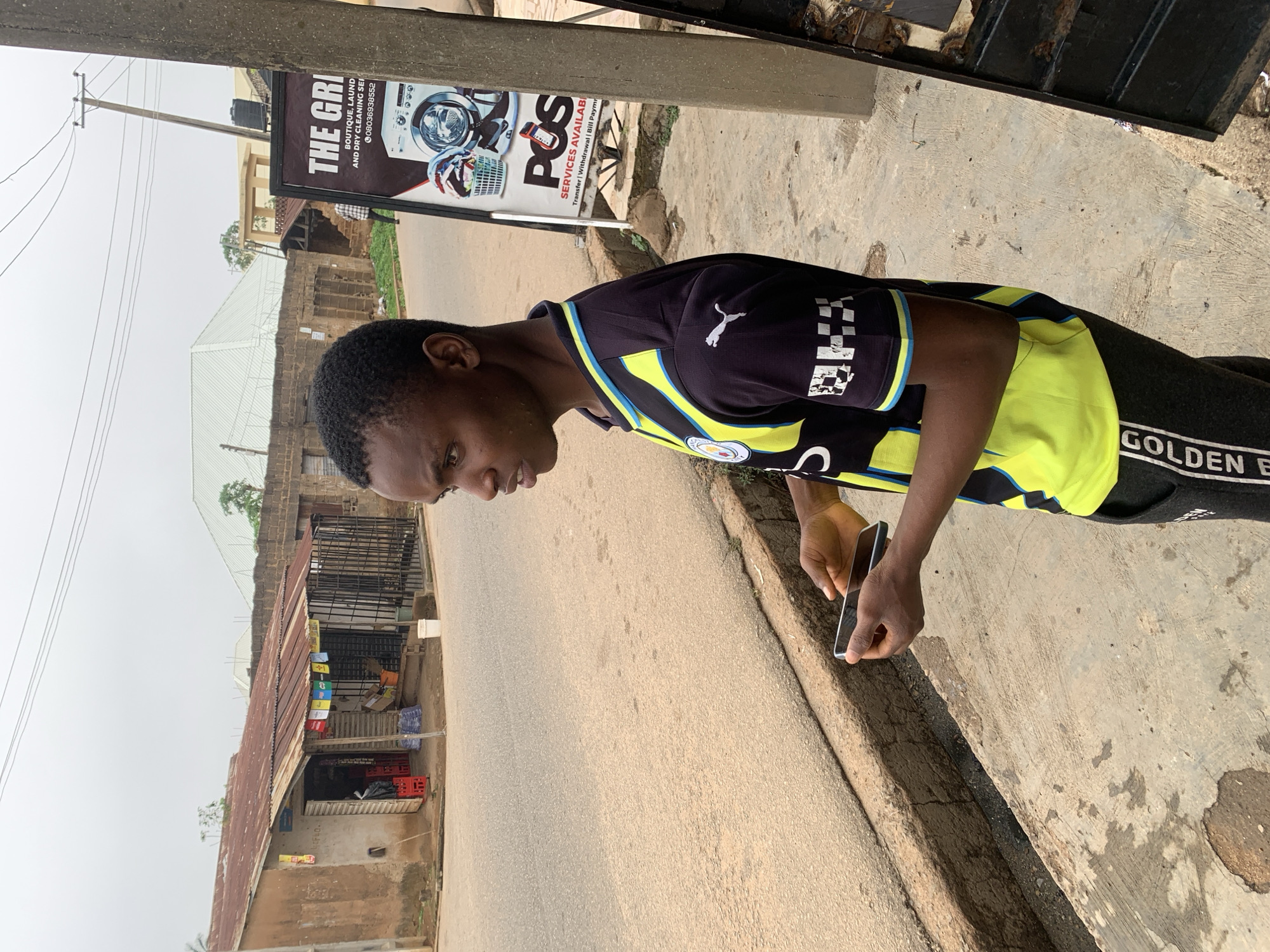 A young Black male stands on a concrete sidewalk next to a paved road in Gura Top, Nigeria. He is wearing a black and yellow athletic jersey and dark pants with white lettering. The male is holding a dark-colored smartphone in his hands, looking to his left. Behind him, a small, rustic shop with a corrugated metal roof is visible, displaying red crates and plastic water bottles. To his right, a tall pole with electrical wires stands beside a sign advertising "THE GRI" services, including laundry, dry cleaning, and POS transactions. The street is mostly empty, with a few distant figures and buildings in the background. The sky is overcast, suggesting a cloudy day.