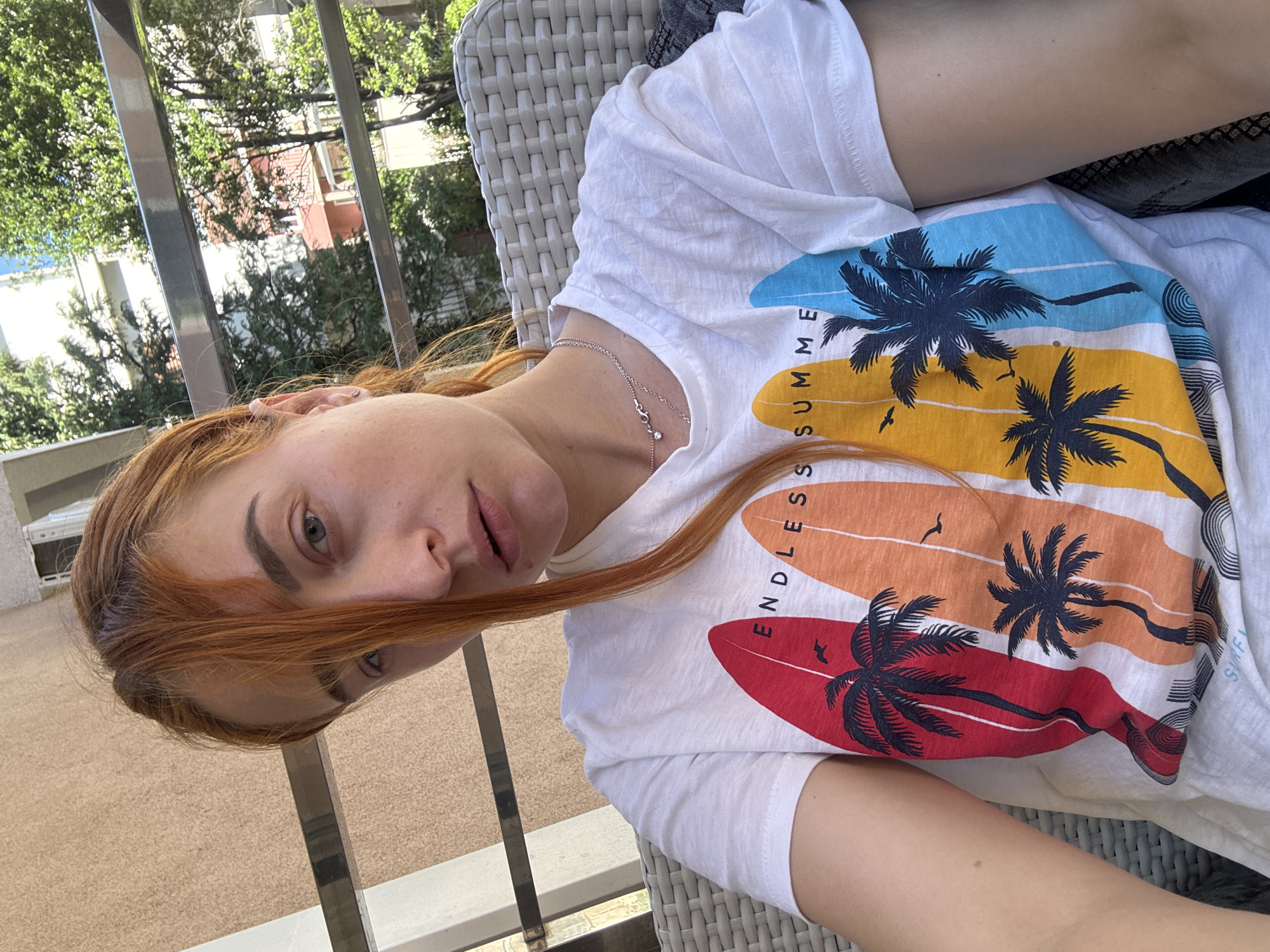 A light-skinned woman with red hair is reclined in a white wicker chair on a balcony. The background shows trees and a red-roofed building. She is wearing a white t-shirt with colorful images of surfboards with palm trees. The text "ENDLESS SUMMER" is visible on the t-shirt. She is wearing a silver necklace. The scene likely takes place outdoors. A metal railing surrounds the balcony. The location is likely in Budva, Montenegro.