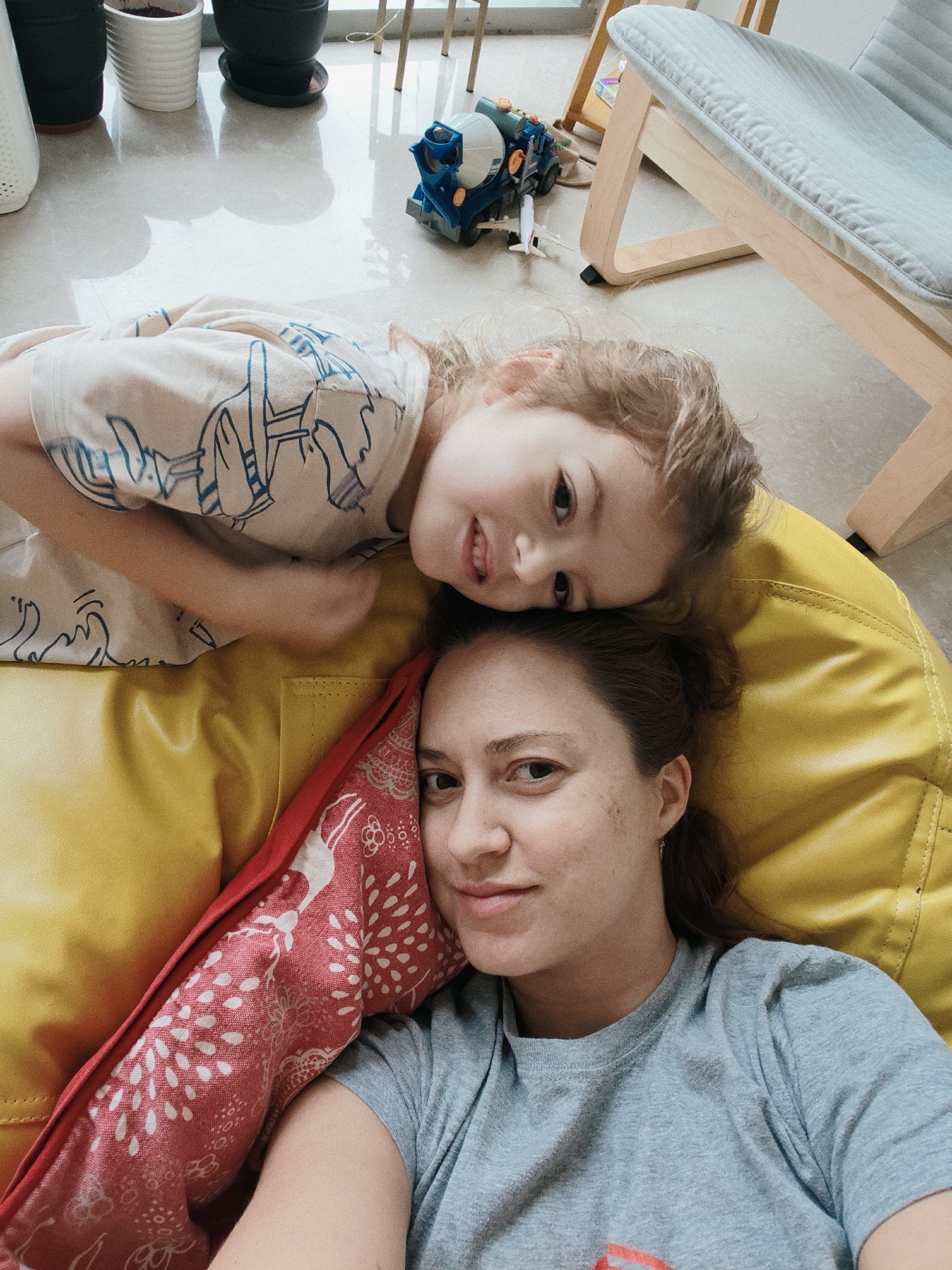 In a residential setting in Mumbai, India, a mother and her young child are lying on a bright yellow beanbag chair. The mother, on the bottom, is wearing a grey t-shirt. Her head rests on the beanbag, and her face is partially visible. The child, on top, has their head resting on the mother's head. The child is wearing a light-colored t-shirt with blue drawings. A red pillow with white floral patterns is nestled between the mother and the child. In the background, out of focus, are several potted plants and a piece of furniture, possibly a chair. A toy truck and a toy airplane are on the floor nearby. The lighting suggests it is daytime, with natural light streaming in from a window.