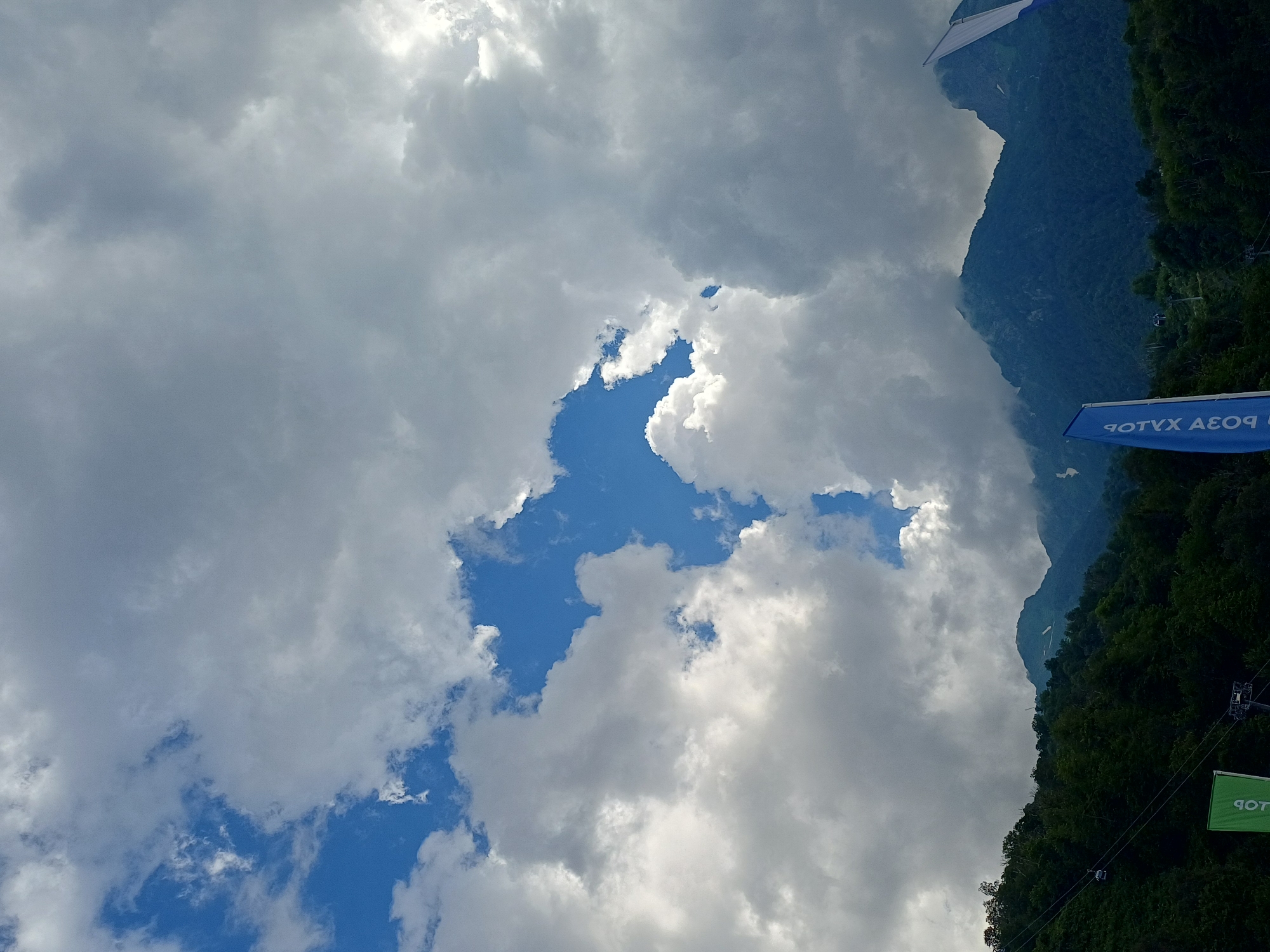 The image captures a dramatic sky over a mountainous landscape in Estosadok, Russia. Dense, gray clouds dominate the upper portion of the frame, with a few patches of bright blue sky peeking through, creating a dynamic contrast. The sun is partially obscured, casting a diffused light. Below the clouds, lush green forested slopes rise, leading to rugged, dark blue mountain peaks. Some of these peaks are shrouded in mist.
In the foreground and midground, there are signs of human activity. A cable car system is visible, with a car ascending a slope on thin wires. A bright green flag with some text is also present, partially obscured. To the right, a blue flag with white lettering, written in reverse, is prominently displayed. The overall impression is of a vast natural landscape with hints of outdoor recreation or tourism.