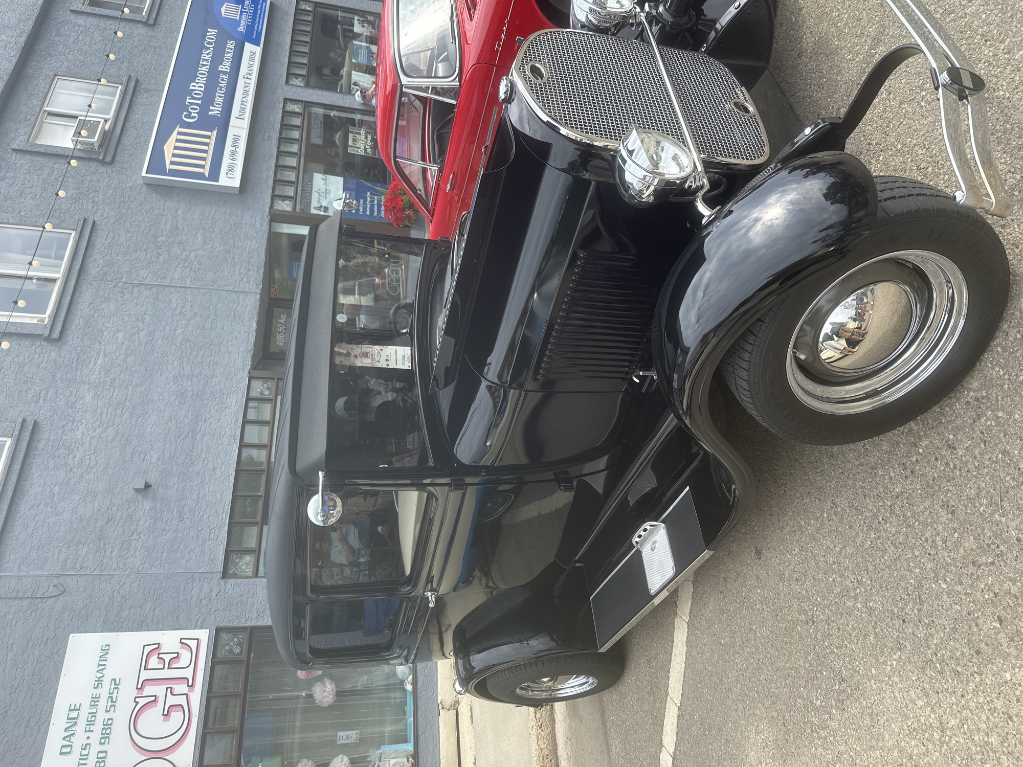 This media file captures a street view featuring a classic black car parked next to a red vintage car. The black car, likely from the 1930s, is the primary focus, showcasing its gleaming chrome accents, prominent grille, and polished black bodywork. The red car is visible behind it, also appearing to be a vintage model.
The background reveals a building with a grey textured facade. Signage on the building indicates commercial businesses, including "GoToBrokers.com Mortgage Brokers" with a phone number and "Dominion Lending Centres." Another sign below announces "DANCE TICS. FIGURE SKATING" with a phone number. The street appears to be paved, with white lane markings visible.
The scene suggests a daytime setting, with natural light illuminating the vehicles and buildings. The overall atmosphere is one of a casual outdoor gathering or event, possibly a car show or a local street event in the city of Leduc, Canada, as hinted by the address "4816-50 Ave." and the business names. No specific activity or event is explicitly depicted, beyond the presence of the cars and businesses. The reflections in the car's chrome and paint suggest a clear sky with some clouds.