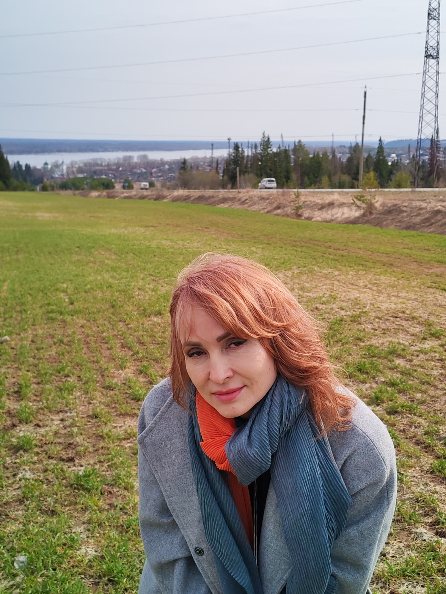 A woman with auburn hair stands in a field of green grass. She is wearing a grey coat and a blue and orange scarf. Behind her is a road, some trees, and a body of water. Power lines and a lattice tower are visible in the distance. The location is Нечкино, Russia.