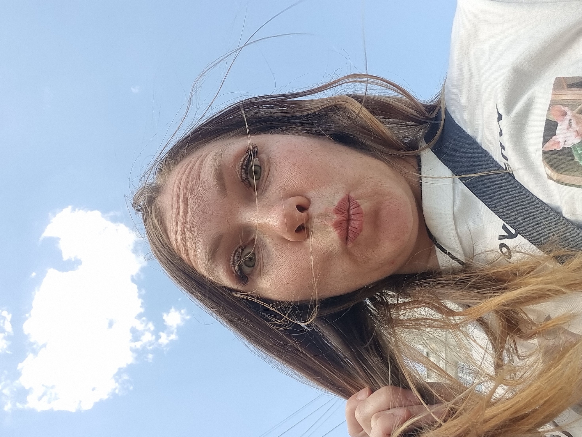 A woman with brown hair and green eyes poses for a selfie against a blue sky with scattered clouds. She is wearing a white shirt with a black and white striped strap visible. She is pursing her lips. The location is Chaikovsky, Russia.