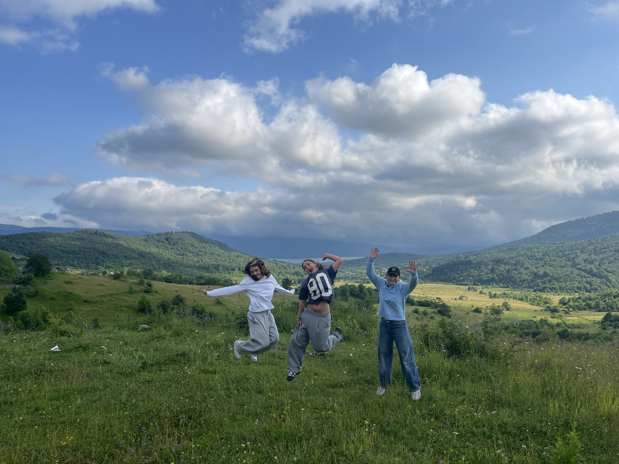 Three individuals are captured mid-air in a playful jump within a verdant, hilly landscape, under a sky adorned with dramatic clouds. The person on the left wears a white long-sleeved shirt and grey sweatpants, with their arms outstretched. The central figure is dressed in a dark t-shirt with the number "80" and grey, baggy sweatpants, striking a dynamic pose. The individual on the right is in a light blue hoodie and blue jeans, waving with both hands. The foreground is a meadow filled with wildflowers and tall grass. In the background, rolling green hills rise towards a distant body of water and more undulating, tree-covered mountains under a bright, cloudy sky. The general location is suggestive of a rural or mountainous region.