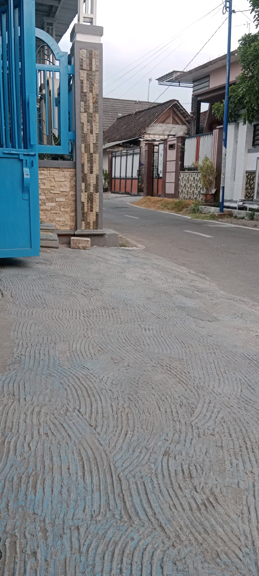 The image depicts a section of a residential street in Soko, Indonesia, focusing on the textured concrete pavement in the foreground and a gate and building in the background. The pavement is light gray with a textured, wavy pattern and some visible blue paint. To the left, a bright blue metal gate is partially open, revealing glimpses of what appears to be fencing or another gate behind it. A concrete pillar adorned with stone tiles stands to the right of the blue gate. Beyond the gate and pillar, a road curves away, lined with houses and fences. One house has a brown tiled roof and a gate with white and orange bars. Another building on the right is white with dark trim and windows. Utility lines are visible against a cloudy sky. The scene is static and appears to be taken during daylight. There are no people or animals visible in the image.