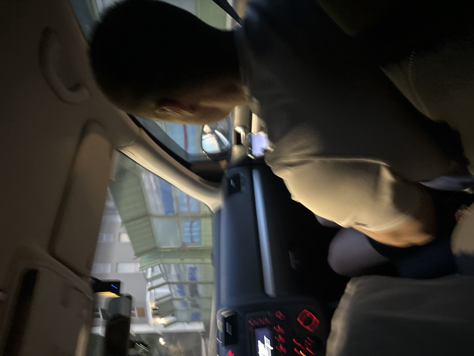 The image shows the interior of a car at night. The driver, a person with short dark hair and wearing a grey t-shirt, is in the right seat, facing forward. Their left leg is visible, clad in dark shorts. A smartphone is held in their right hand, illuminating their face with its screen. Outside the car, a building with multiple windows and a green, grated fence are visible. The dashboard of the car is dark, with red illuminated buttons and a display screen. A dashcam is mounted on the windshield. The environment suggests the car is in motion, likely on a road within the town of Rafailovići, Montenegro.