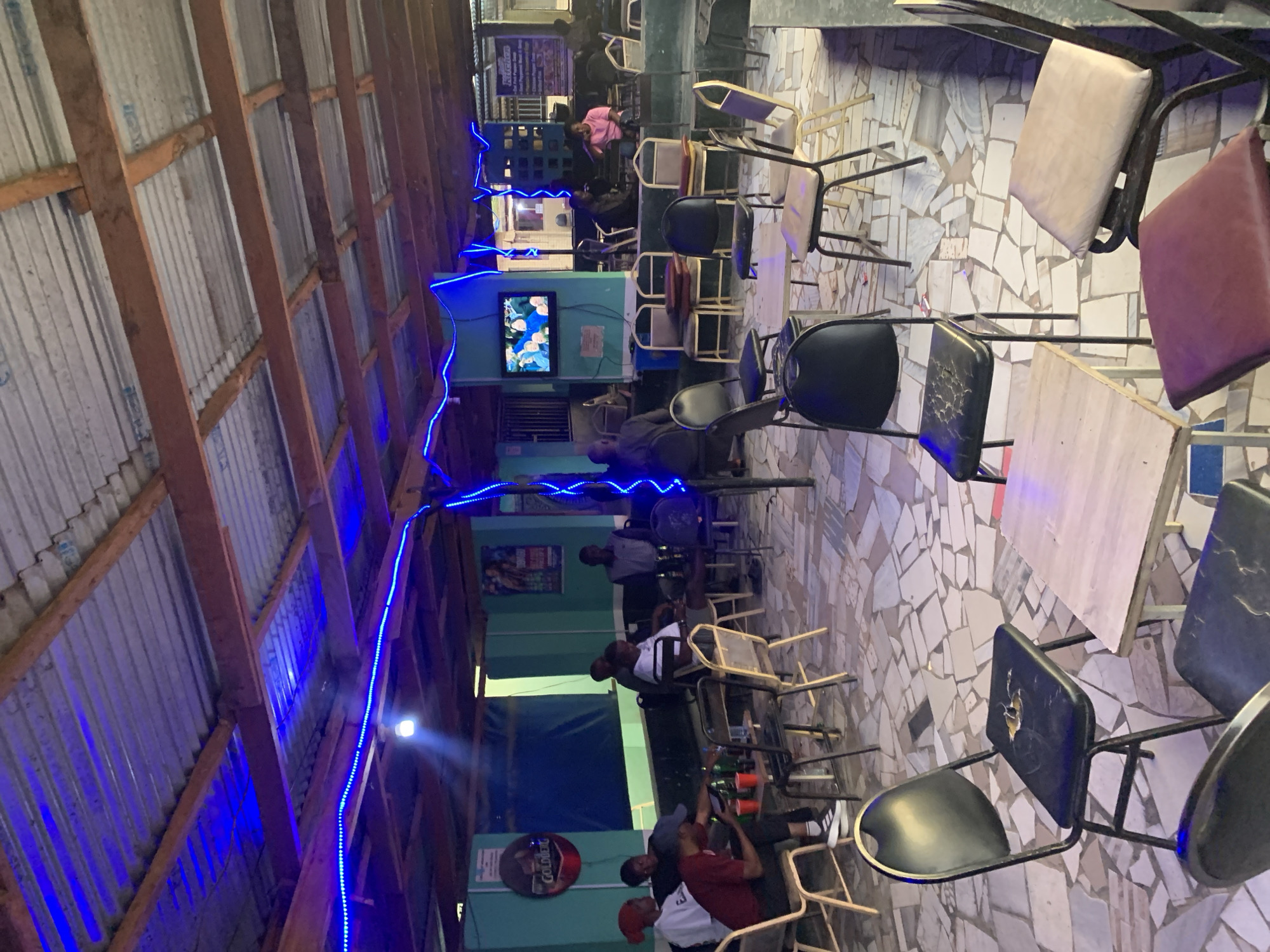 The image depicts the interior of a bar/restaurant in New Stadium, Nigeria. Patrons are seated at tables, some are drinking and socializing. A television screen displays a program. The venue has a corrugated metal roof with exposed wooden beams illuminated by blue LED strip lights. The floor is tiled with irregular white and grey pieces. The furniture includes metal-framed chairs with various colored cushions. There are beer bottles and plastic cups on some tables. A round sign for "Goldberg" beer is visible on one of the walls.