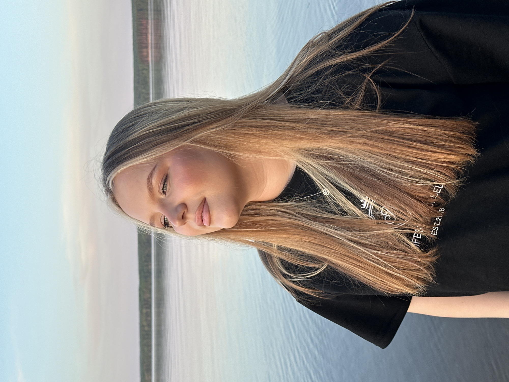 A woman is posing for a portrait. She has long blonde hair, fair skin, and is wearing a black shirt with white writing on it. She is standing outside, possibly near the city of Chaikovsky, Russia as inferred from the landscape, which appears to be near a body of water such as a lake or river. The water reflects the sky, which appears to be light blue, possibly around sunrise or sunset. Trees can be seen in the distance on the opposite bank.