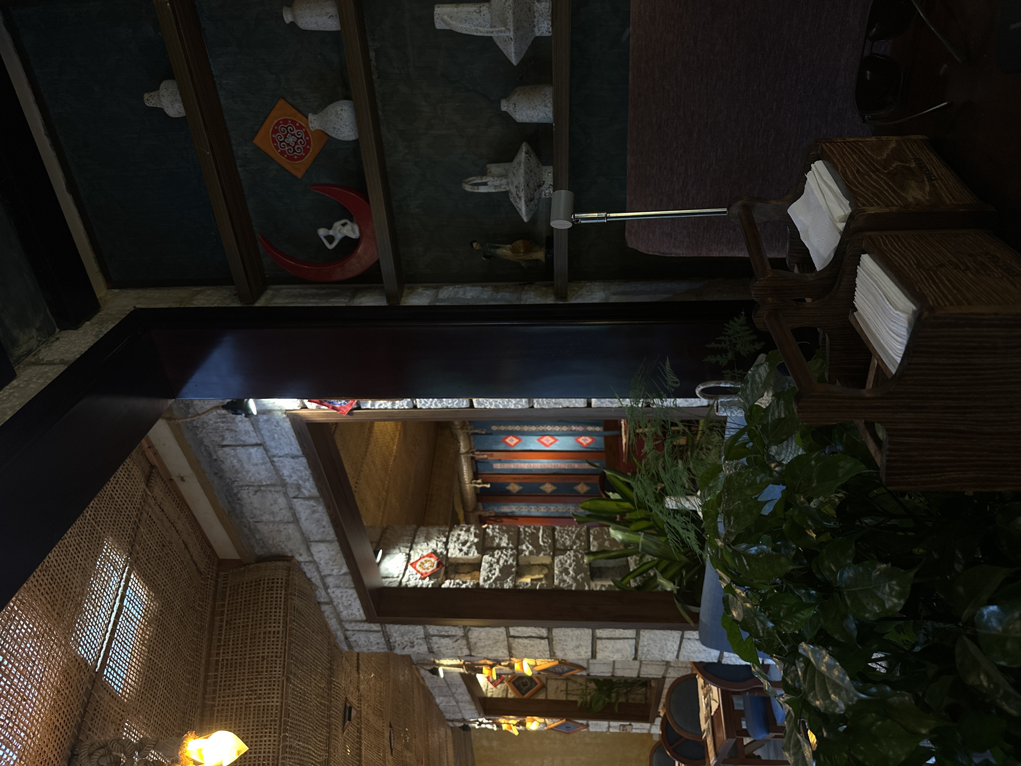 The image, rotated 90 degrees, depicts a partially obscured interior scene. Architectural details include stone and brick walls, woven reed-like panels, and decorative ceiling elements featuring what appears to be crafted objects and shapes.
In the foreground, a wooden napkin holder is visible. Further back, plants are prominent. The architectural style suggests a cultural or traditional aesthetic.
The setting suggests a location within a building in the Sa Pa Ward, Vietnam. Details such as the construction materials, room lighting, and decorative elements indicate an interior design that is specific to the region.