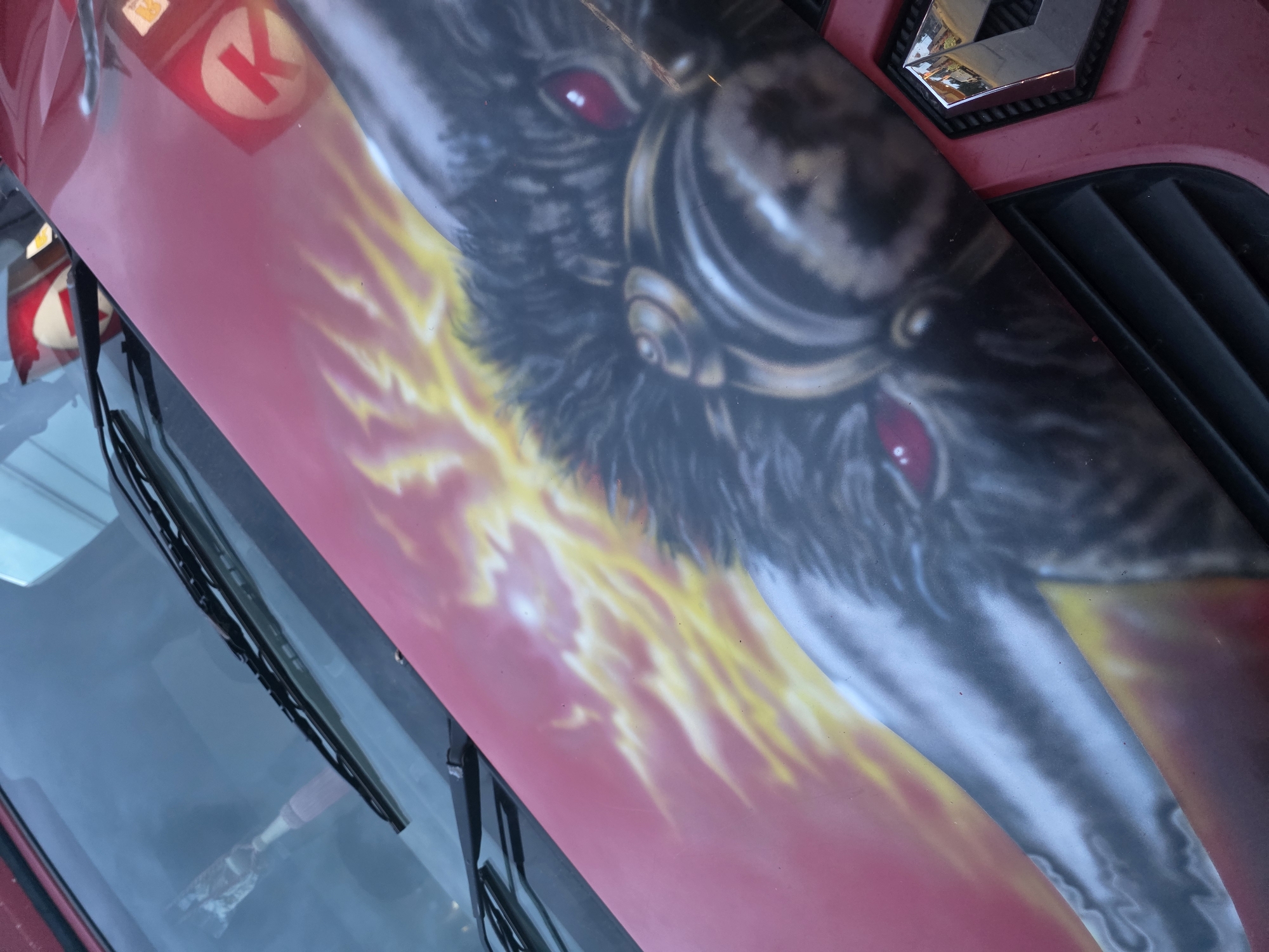 The image displays a close-up, angled view of the front hood and lower windshield of a red vehicle, likely a car or a van, in the city of Unknown, Unknown. The vehicle's hood features custom airbrushed artwork depicting a dark, furry beast's head with piercing red eyes and metallic accents around its muzzle/neck area. Behind the beast, a vibrant yellow and orange flame-like pattern extends across the red surface of the hood.
The windshield, visible at the top, is angled, with black windshield wipers resting horizontally. Reflections are prominent on both the windshield and the vehicle's painted surfaces. On the windshield, reflections include what appears to be a light blue wall or sky and a blurred, elongated reddish object, possibly a banner or flag. On the red hood, two distinct reflections of a circular logo with a white "K" inside a red circle are visible, consistent with a Circle K convenience store sign.
The lower right portion of the image shows part of the vehicle's front grille, which is black plastic with horizontal slats. A silver, chrome-finished Renault emblem is affixed to the red bodywork just above the grille, reflecting distorted images of the surroundings. The overall paint surface, particularly the red areas, shows minor dust or debris.