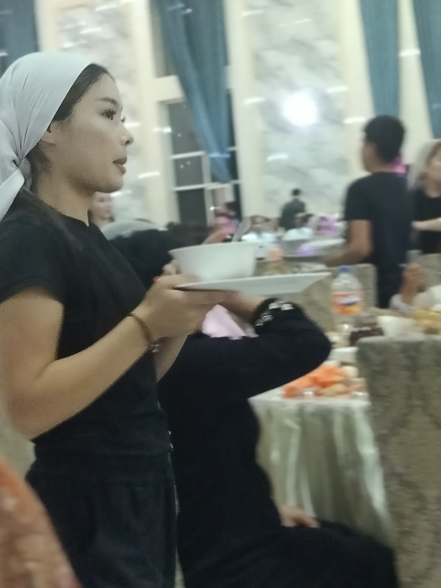 In a dimly lit, indoor setting, possibly a banquet hall in Ақсуабат, Kazakhstan, a young woman in a light-colored headscarf and a black t-shirt is visible from the side. She is holding a white bowl and plate, and a beaded bracelet adorns her wrist. She appears to be serving or presenting the items. In the background, other individuals are blurred, with one person wearing a black t-shirt and another in a black head covering. Tables laden with food are also partially visible, suggesting a meal or event is taking place. The environment features large, possibly marble-patterned columns and blue drapes.
