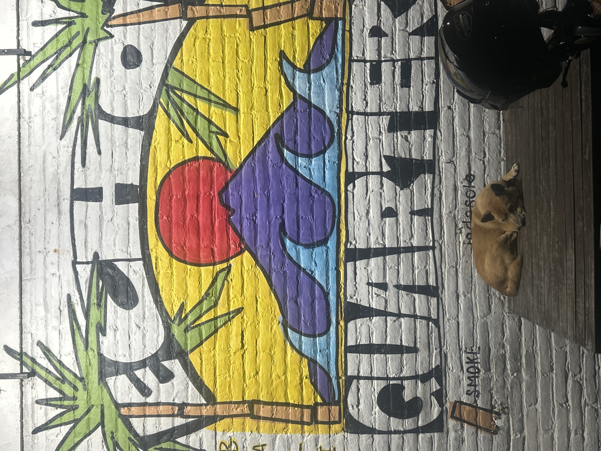 The image shows a mural on a brick wall in North Kuta, Indonesia. The mural depicts a stylized scene with palm trees, a red sun, and a purple and blue shape against a yellow background. The word "GUA" is painted vertically on the right side of the mural. In the lower right corner of the image, a small brown dog is lying on the ground near the wall. Above the dog, a black motorcycle helmet is visible. "Smoke" is also written in a dark color, right below the word "Gua".