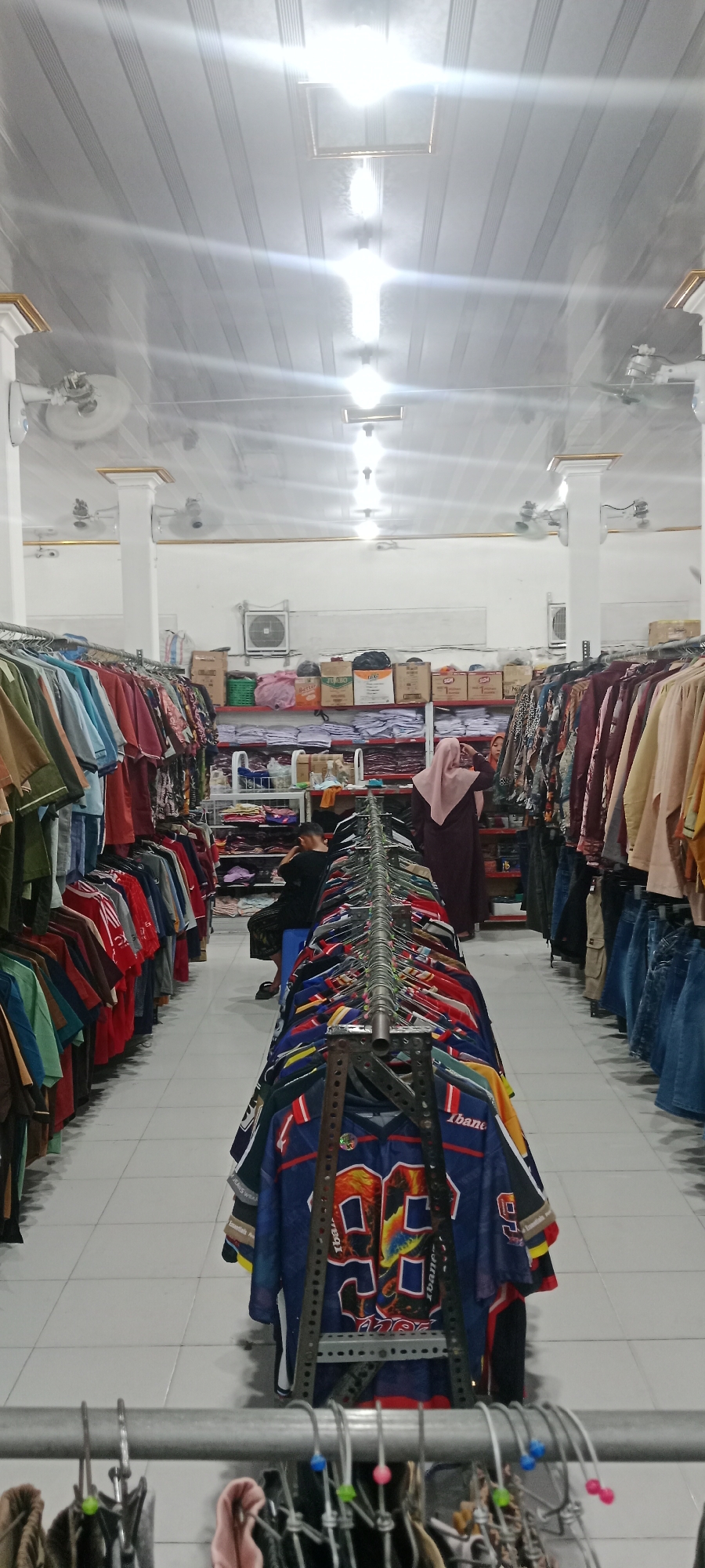 The image depicts the interior of a clothing store in Rengel, Indonesia. The perspective is from within an aisle, looking down the length of the store. Clothes are displayed on racks lining both sides of the aisle and in the center. The ceiling is white with a metallic sheen and features rows of fluorescent lighting and ceiling fans. The walls are also white.
On the left side of the aisle, clothing racks display a variety of garments, including what appear to be long-sleeved shirts and dresses in various colors and patterns, predominantly in muted tones and earth tones.
On the right side of the aisle, similar clothing racks are filled with garments. Towards the back, a woman wearing a light pink hijab and a dark maroon abaya is standing. Another person, dressed in a black shirt and pants, is seated on a blue stool, looking towards the shelves.
In the center of the aisle, a long rack of t-shirts is prominently displayed. These t-shirts feature bold designs, including what appears to be sports jerseys with large numbers, such as "99", and brand names like "ibane".
Further down the store, shelves are visible stocked with folded clothing, possibly uniforms or t-shirts, and boxes. Some of these boxes are labeled "JUMBO".
The floor is tiled with white square tiles. The overall impression is of a well-lit, modestly stocked retail environment.