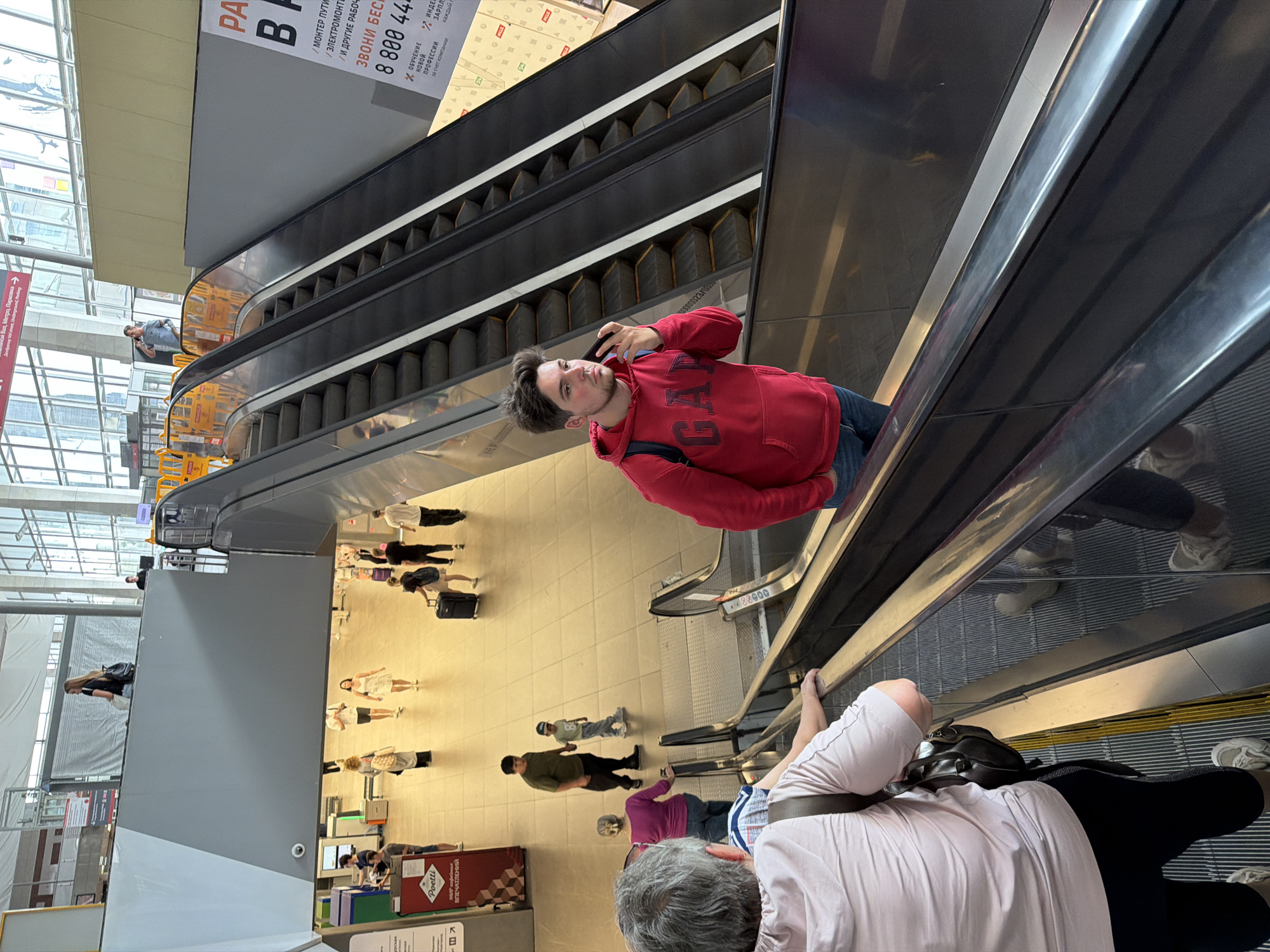 This is a vertically oriented, eye-level shot taken in an indoor public space, likely a metro station or large transit hub in Moscow, Russia. The primary focus is an escalator with several people on it and around it.
In the foreground, a person wearing a light pink shirt and dark pants is seated on the escalator, looking down. Behind them, the escalator steps are visible, with other individuals ascending or descending. The most prominent subject is a young man wearing a red GAP hoodie and blue jeans, taking a selfie with his phone as he rides the escalator upwards. He has brown hair and appears to be looking at his phone. His red hoodie is bright and stands out against the metallic tones of the escalator.
The background reveals a bright, airy space with large windows and a high ceiling. There are directional signs in Russian, including one that says "Метро Парковка" (Metro Parking) and "ВПЕЧАТЛЕНИЙ" (Impressions). Another sign displays a phone number and advertising for training. People are seen walking on the tiled floor below the escalator, some with luggage, suggesting travel. The scene appears to be during the daytime due to the ample natural light filtering in. The overall atmosphere seems to be one of transit and daily activity.