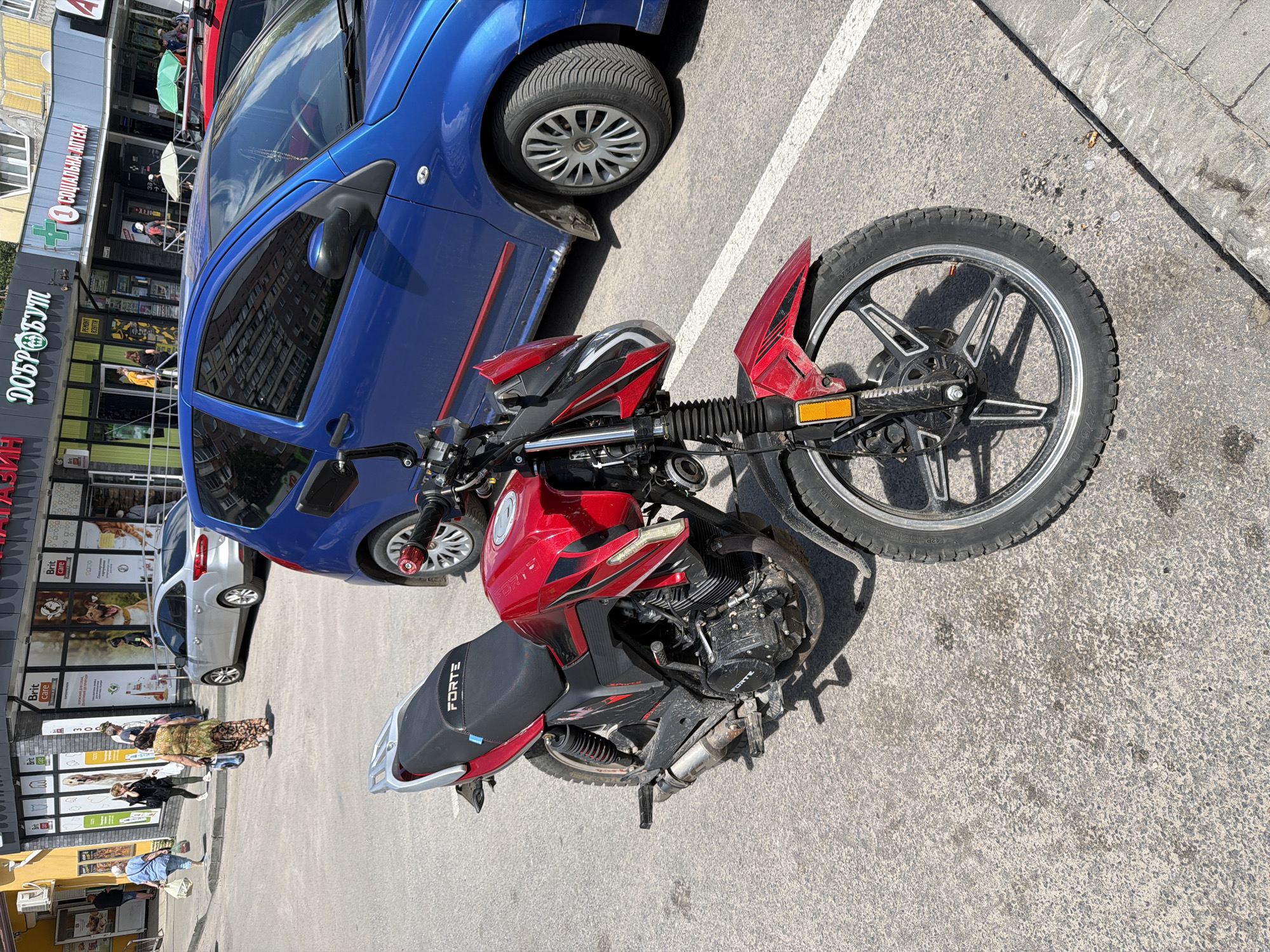 The image depicts a red motorcycle parked on an asphalt surface in front of a building with storefronts. The building has a yellow facade on the left and appears to be a commercial establishment with signage in Cyrillic. A silver sedan is parked to the left of the motorcycle, and a blue hatchback is parked to the right, partially obscuring the motorcycle's rear. Several pedestrians are visible in the background, walking on a sidewalk in front of the stores. The environment suggests an urban setting, likely a street in Dnipro, Ukraine, given the Cyrillic signage. The motorcycle is the primary subject, positioned in the foreground and taking up a significant portion of the frame. The scene is captured during daylight, with clear shadows indicating a sunny day.