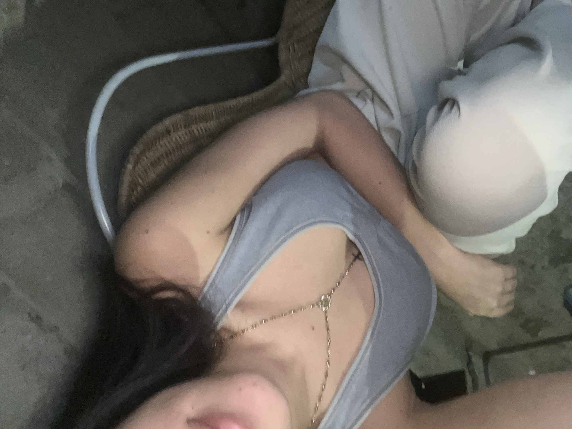 The image shows a close-up, high-angle view of a person sitting on a wicker chair in an outdoor setting, possibly a patio or garden area. The person, whose face is partially visible, is wearing a light blue tank top and light-colored pants. A gold necklace with a square pendant is visible around their neck. The background is dark and out of focus, with hints of what might be concrete or rough ground. There are no discernible landmarks or architectural features that would suggest a specific location within Juan Griego, Venezuela. The lighting appears to be natural and dim, possibly from dusk or an overcast day.