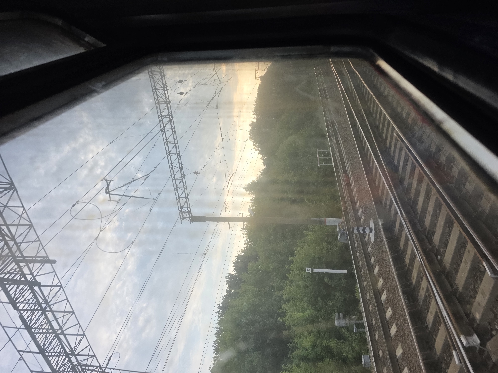 A view from a train window shows a set of parallel train tracks stretching into the distance, bordered by a dense green forest. Overhead, intricate power lines and supporting structures crisscross the sky, which is illuminated by a soft, hazy sunset. The light filters through the trees and the window, creating a slightly diffused and ethereal atmosphere. The window itself appears somewhat dirty, with smudges and streaks visible, especially in the upper portion. A few signal posts and markers are visible along the tracks, indicating the railway's operational status. The overall impression is one of travel and transition, with the natural landscape providing a backdrop to the industrial infrastructure of the railway.