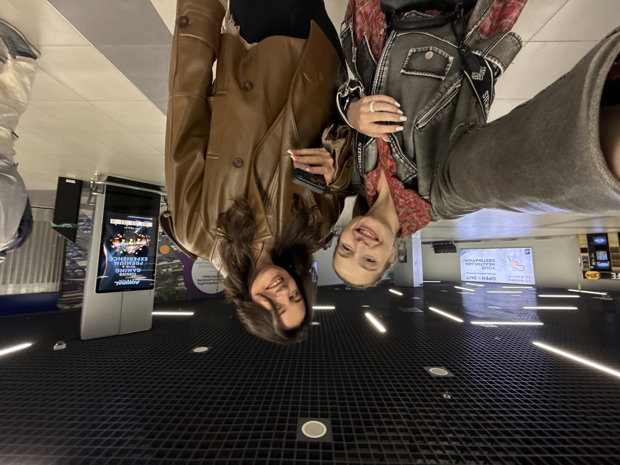 Two women are smiling in an upside-down selfie. The woman on the left is wearing a brown leather-look jacket and holding a dark-colored phone in her right hand. The woman on the right is wearing a grey leather-look jacket over a red plaid shirt. The setting appears to be inside, possibly in a commercial or transportation hub, indicated by the illuminated patterned floor, advertisements, and a digital display. The location could be in the city of Surcin Urban Municipality, Serbia.