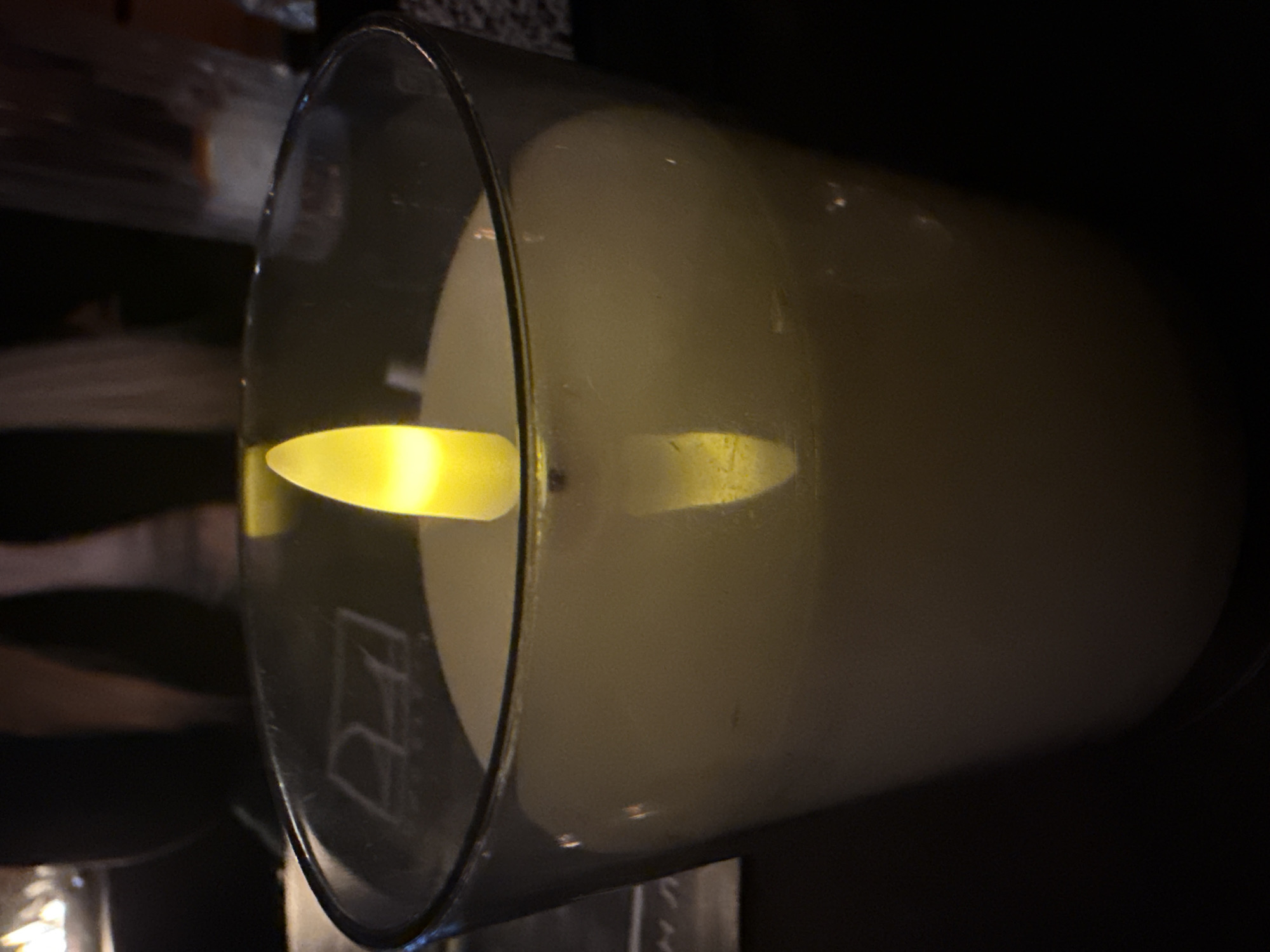 A close-up, vertically oriented photograph depicts a white LED candle contained within a clear, tapered glass holder, positioned on a dark surface in a dimly lit indoor environment. The artificial, yellow-orange flame-like element of the candle emits light, casting a reflection on the candle's white, wax-like body and the inner surface of the glass. The background is out of focus. To the immediate left, a faint, reflective, stylized white logo, resembling "D'angelo" or "DA", is discernible on a dark surface, possibly a menu or tabletop. To the right, an out-of-focus, translucent drinking glass with indistinct contents is visible. Beyond these objects, blurred human figures are present in the dark background, with one appearing to wear dark clothing. The scene's ambient lighting is primarily derived from the candle. Based on the visible logo, the location is identified as D'angelo Pizza & Pasta in Varna, Bulgaria.