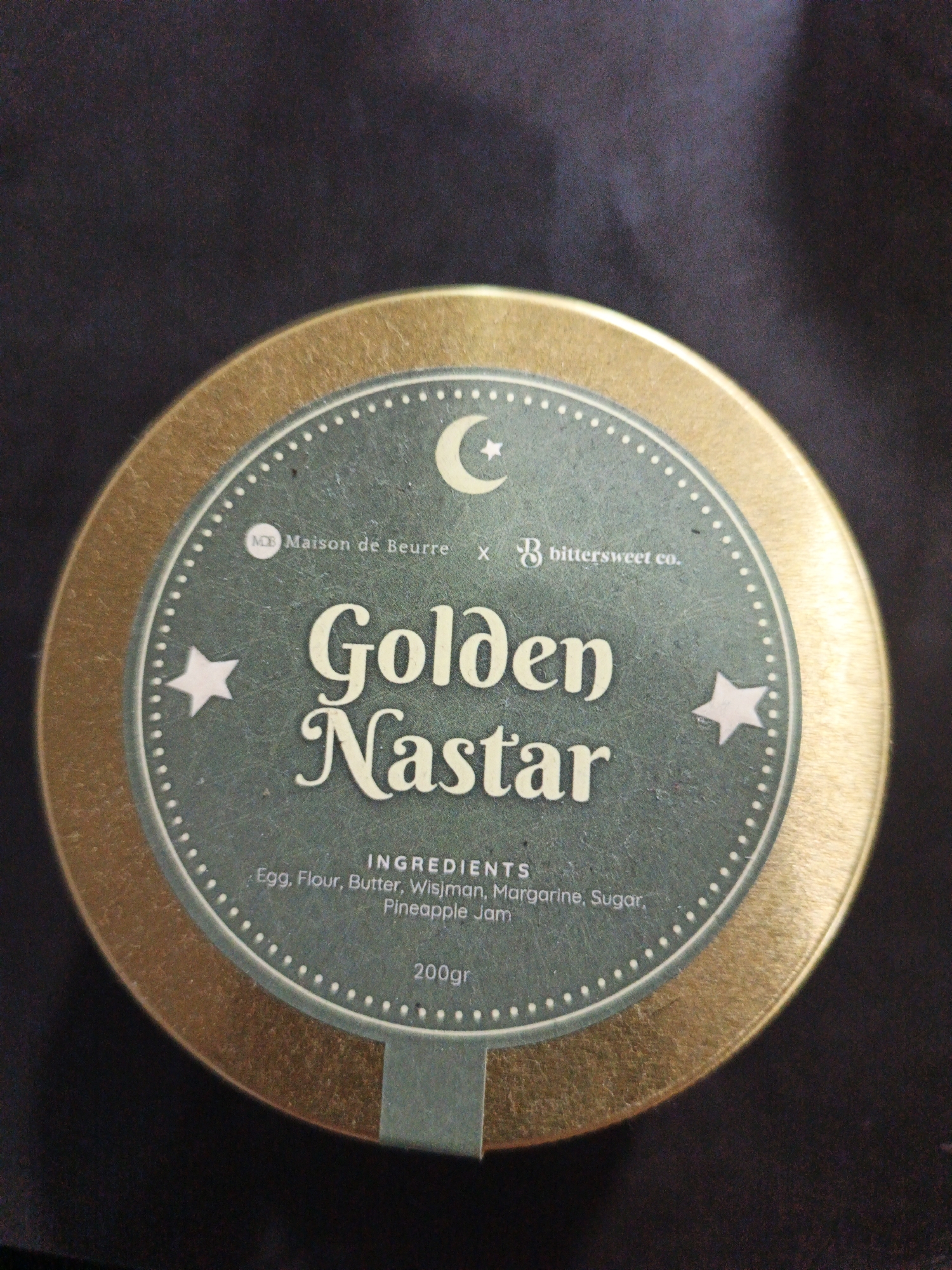The image shows a close-up of a round, golden tin with a dark background. The tin has a teal-colored label in the center with white text and graphics. The text "Maison de Beurre" appears above a crescent moon graphic, and "bittersweet co." appears to the right. Below this, the words "Golden Nastar" are written in large, decorative font. Further down, "INGREDIENTS" is written in smaller text, followed by a list of ingredients: "Egg, Flour, Butter, Wisjman, Margarine, Sugar, Pineapple Jam". At the bottom of the label, "200gr" is displayed. The tin is positioned on a dark, textured surface. There are no people or discernible environmental features in the image that would indicate a specific location in Kelapapati, Indonesia.