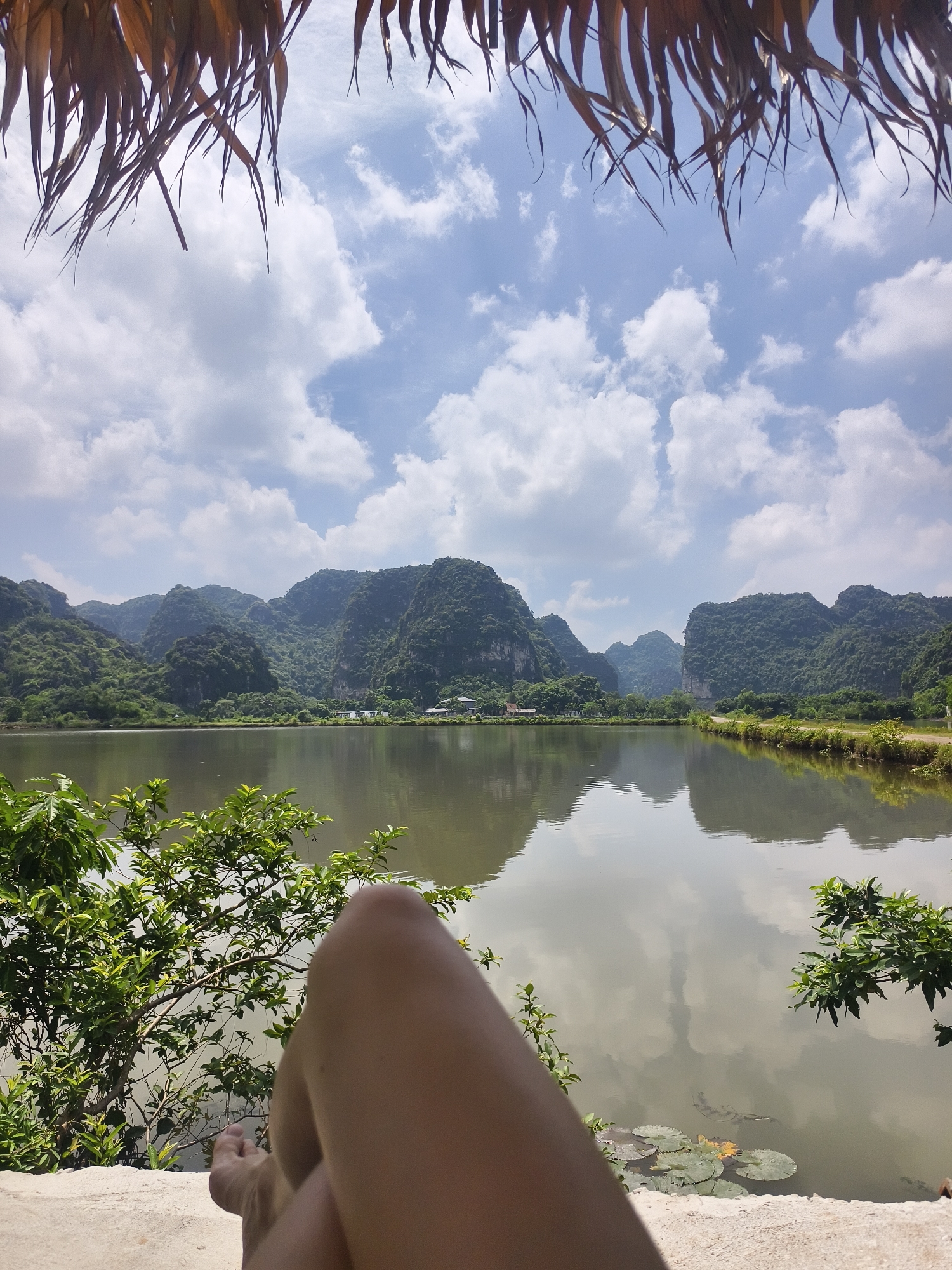 A person's bare legs and feet are visible in the foreground, resting on a light-colored surface. They are looking out at a tranquil lake, bordered by lush green mountains in the distance. The sky above is a mix of blue and white clouds, with the fronds of a thatched roof overhead partially obscuring the view. The lake reflects the surrounding landscape and the sky. In the water, lily pads are visible. The scene appears to be set in Hoa Lư Ward, Vietnam, known for its karst landscapes and waterways.