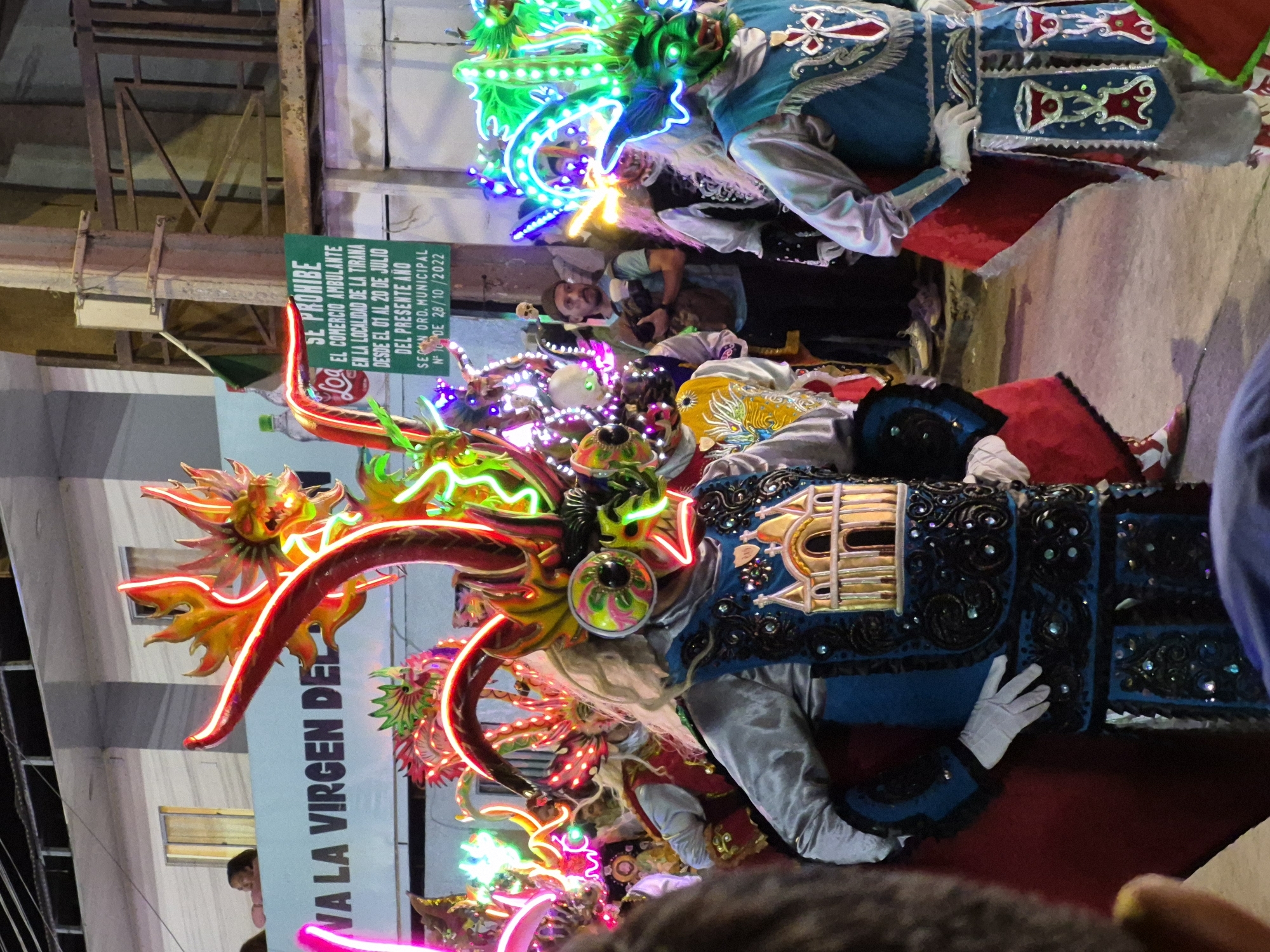 The image captures a nighttime street scene in Pozo Almonte, Chile, featuring a vibrant parade or festival. The foreground is dominated by individuals in elaborate, brightly lit costumes, primarily featuring dragon or monster-like masks adorned with neon lights in red, green, and yellow. One figure wears a blue and silver costume with a detailed depiction of a building on its chest and white gloves. Another figure in the background wears a similar costume with a red and white striped pattern on their legs. In the background, a sign on a building reads "SE PROHIBE EL COMERCIO AMBULANTE EN LA LOCALIDAD DE LA TIRANA DESDE EL 01 AL 20 DE JULIO DEL PRESENTE AÑO SEGUN ORD. MUNICIPAL N° 100 DE 28/10/2022," indicating a prohibition on street vending during a specific period in La Tirana. People are visible in the background, some observing the event, while one man holds a camera. The environment appears to be an urban street with buildings and structures illuminated by artificial lights, creating a festive and energetic atmosphere.