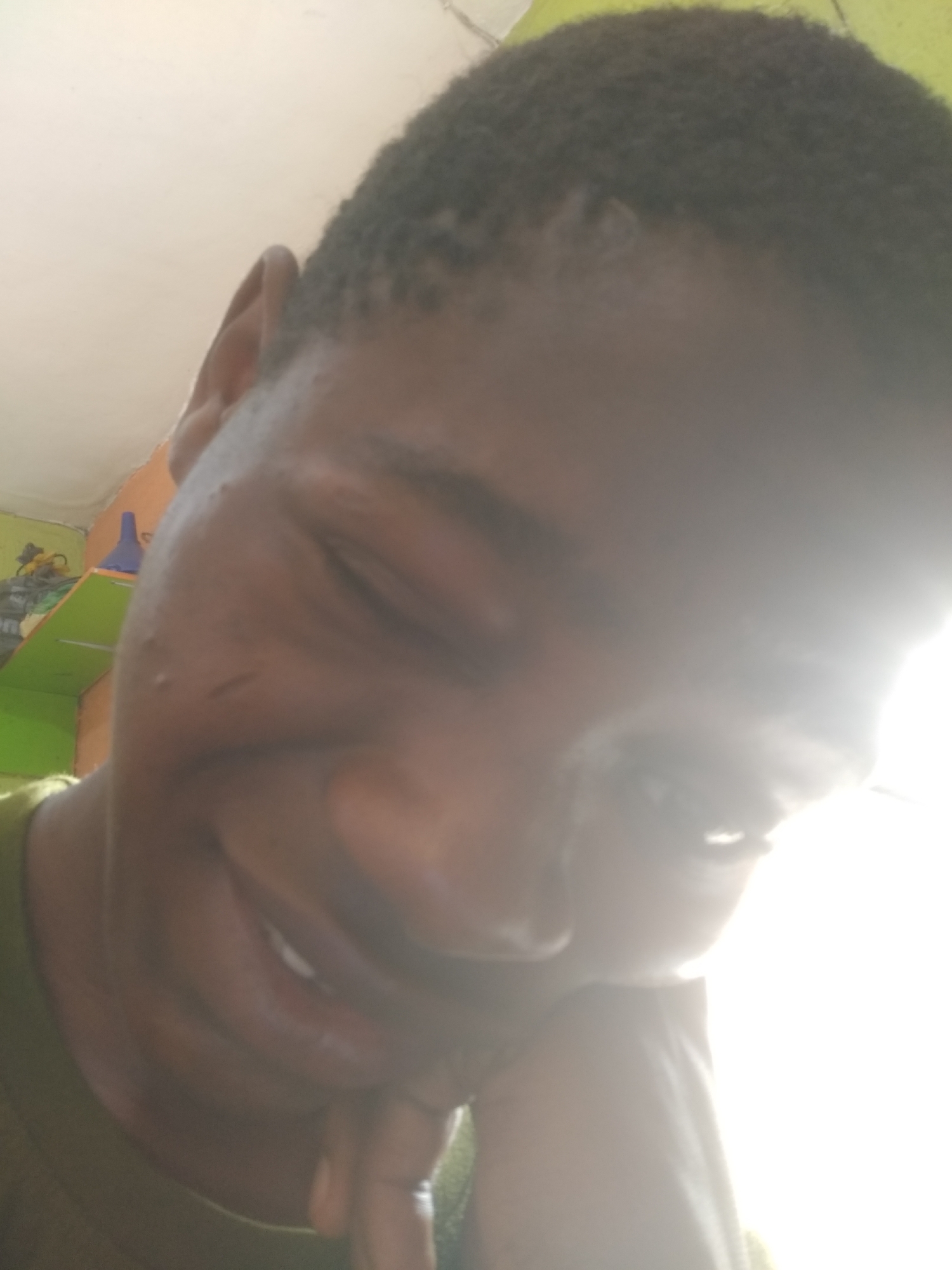 A close-up shot of a person with dark skin, short black hair, and wearing a dark green shirt. The person is smiling with their eyes slightly closed. Their face is angled downward and resting on a hand. The background is a room interior with a shelf visible, containing various items. The location is presumed to be Shangisha, Nigeria.