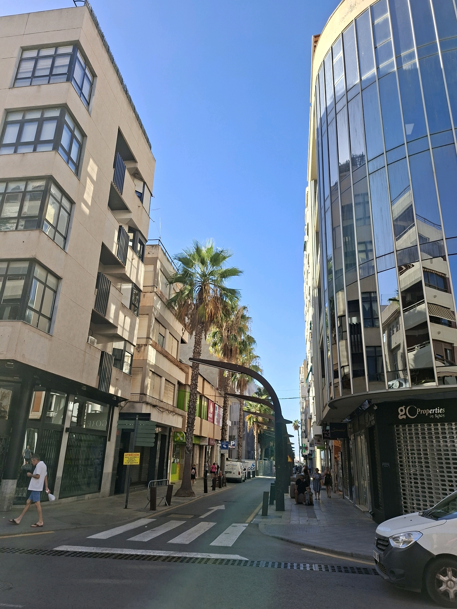 This is a street-level, eye-level, full-shot photograph taken in Torrevieja, Spain. The scene depicts a street lined with buildings and palm trees under a clear blue sky.
On the left side of the street, a multi-story building with a beige facade features numerous windows, some recessed with balconies. The ground floor has large glass storefronts. A person in a white t-shirt and blue shorts is walking on the sidewalk, carrying a white object.
On the right side of the street, a modern building with a curved facade is covered in reflective glass panels, mirroring the sky and surrounding buildings. The ground floor of this building houses businesses with rolling metal security shutters. One storefront displays the logo "GC Properties in Spain".
A pedestrian crossing is visible in the foreground, marked with white stripes. The street itself is paved with asphalt. Several palm trees are interspersed along the sidewalk, with their fronds reaching towards the sky. Other buildings of varying heights and architectural styles are visible further down the street, some with awnings and shops. A few people are scattered along the sidewalk, walking or standing. A white van is parked on the left side of the street in the distance, and a white car is parked on the right. The overall impression is of a sunny day in an urban environment with a Mediterranean feel.