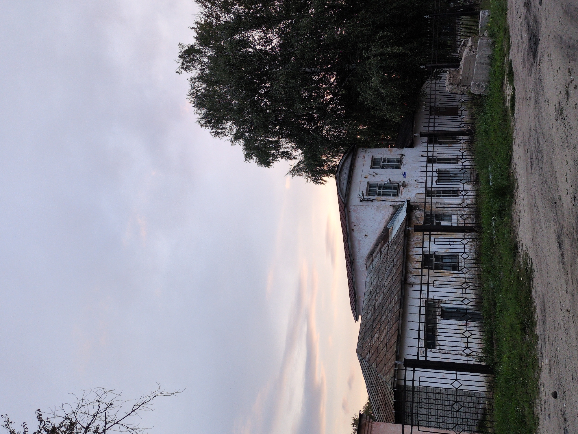 The image depicts an old, weathered, white building with a rusty, corrugated metal roof. A black wrought-iron fence separates the building from a dirt road. To the right of the building, a large, leafy tree dominates the frame. The sky is overcast with hints of pink and orange from what appears to be a sunset. The scene is set in Kulebaki, Russia, suggested by the architectural style and the presence of mature trees, common in the region. There are no people visible in the image. The overall impression is one of a quiet, somewhat neglected, rural or semi-urban setting at dusk.