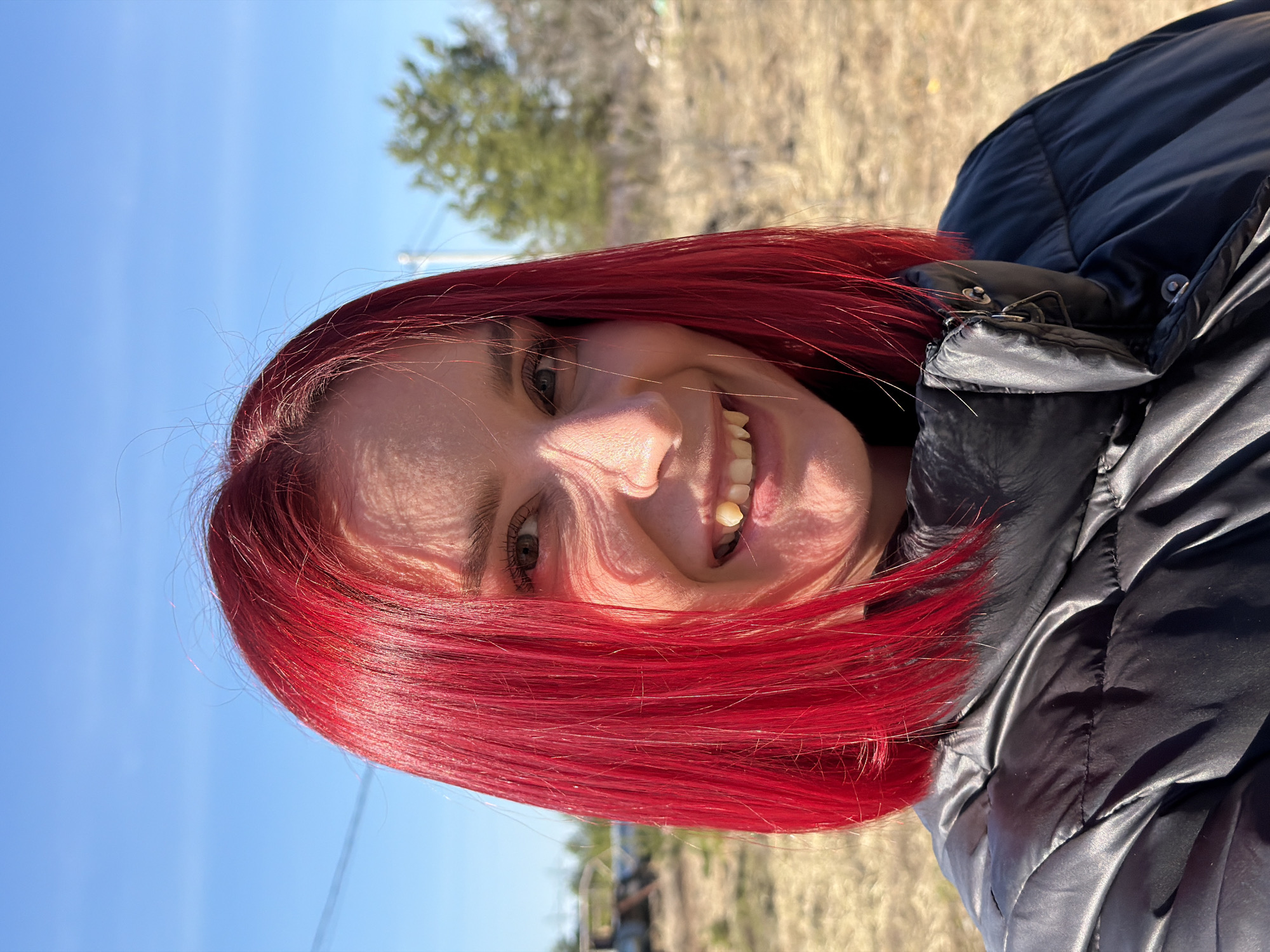 A selfie-style portrait shows a woman with bright red hair smiling broadly. Her hair is cut in a short bob and appears freshly dyed. She has fair skin and visible wrinkles around her eyes. She is wearing a black, shiny puffer jacket. Behind her, the background consists of a clear, blue sky, suggesting a sunny day. There's also a blurry landscape with dry grass and sparse vegetation, including a tree in the distance. Given the context of Chaikovsky, Russia, it is likely an outdoor area within or near the city. Specific locations or landmarks are not discernible in the image.