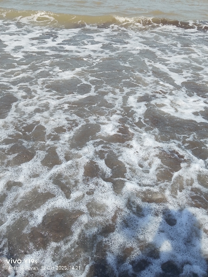 The image displays a detailed, downward-angled view of agitated coastal water. The water exhibits a murky, brownish-grey coloration, characteristic of sediment or reflections, and is extensively covered with white, frothy sea foam and numerous small bubbles, indicative of active wave action and turbulence. Undulating wave patterns are discernible throughout the frame, with more defined crests visible towards the upper portion. No individuals, marine life, or specific man-made objects are visible within the water. A faint, dark, indistinct shadow is cast upon the water's surface in the lower right quadrant. A digital watermark in the bottom left corner reads: "vivo Y19s Akhtar & Arvan, 12 Okt 2025, 14.21". While the scene is consistent with a coastal environment, the image contains no specific visual identifiers to confirm the user-provided location of Limbangan, Indonesia.