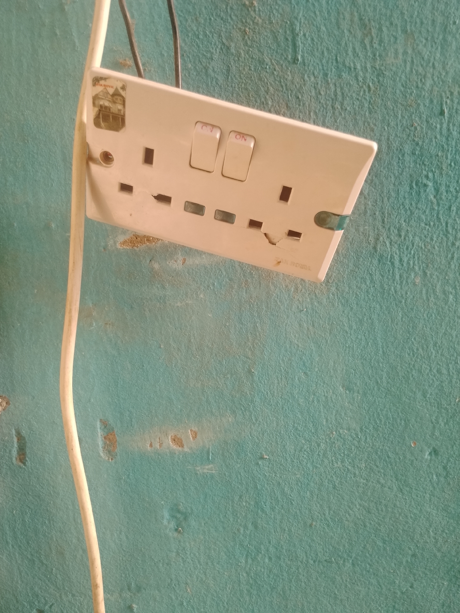 The image shows a close-up, slightly angled view of a white electrical socket mounted on a turquoise, textured wall in Jalingo, Nigeria. A white electrical cord hangs vertically along the left side of the socket. The socket has two sets of three rectangular openings and two individual switches. One switch is labeled "ON". A small, rectangular sticker with an image of a building and the text "ZAK ROYAL" is adhered to the top left of the socket. The wall displays some discoloration and patches of exposed plaster, indicating wear and tear. The lighting suggests an indoor environment, likely a room with a plain wall. No people or animals are visible in the image.