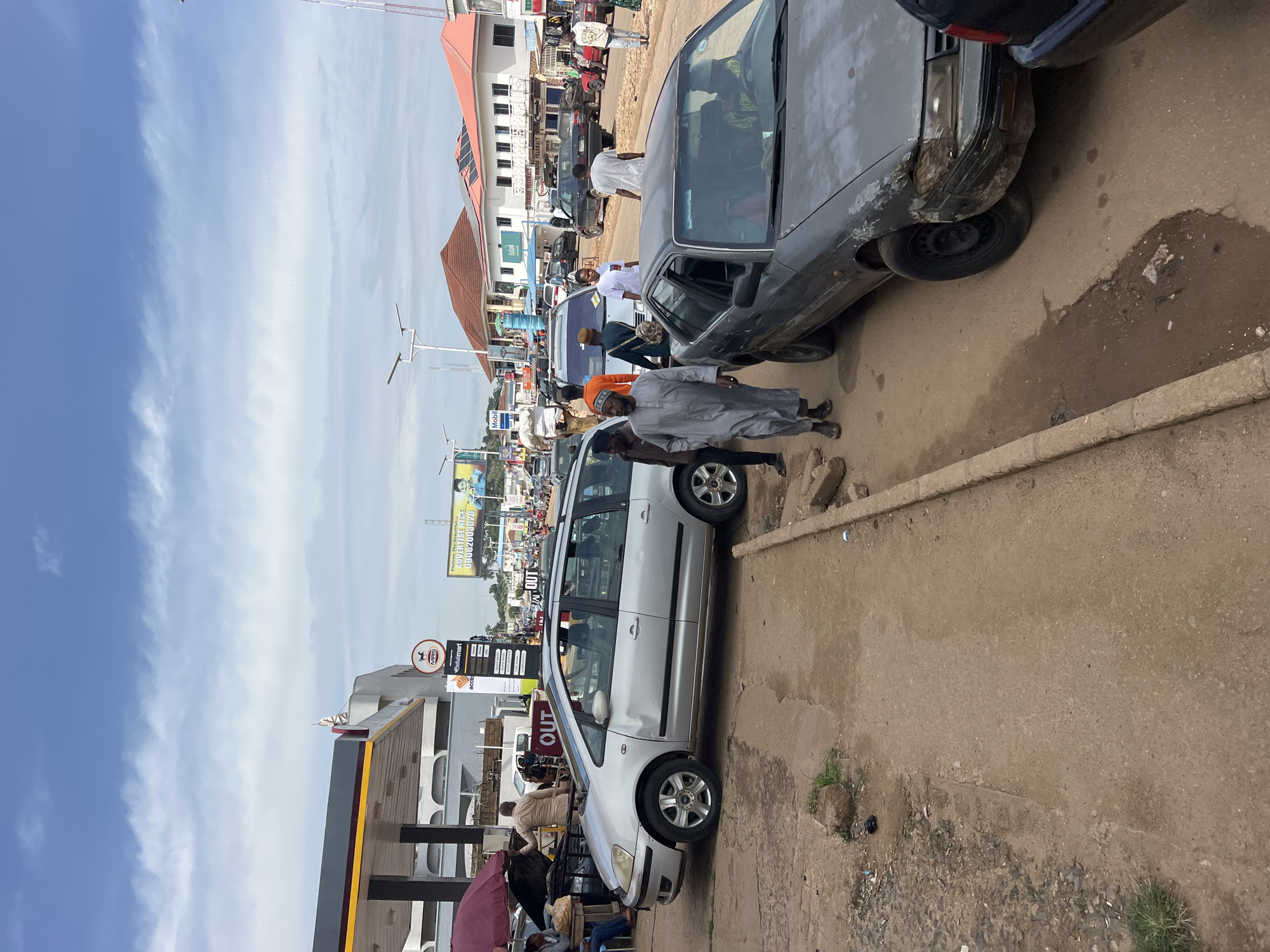 The image depicts a street scene in Jos, Nigeria, under a cloudy sky. In the foreground, a gray sedan with significant damage to its front bumper and hood is parked on the right side of the road. To the left of the sedan, a sidewalk area is visible, with some debris and patchy vegetation. Further left, a silver minivan is parked.
Several people are present in the scene, primarily walking or standing near the vehicles and the roadside. A man in a gray robe and cap is walking towards the foreground on the left side of the road. To his right and further down the street, other individuals in casual attire are visible.
In the background, a gas station with its canopy is prominent on the left. Across the street, a row of buildings lines the road, including what appears to be a pharmacy with red and white signage. Vehicles are seen on the road, indicating traffic. Advertisements are posted on billboards in the distance. The overall environment suggests a busy urban commercial area.